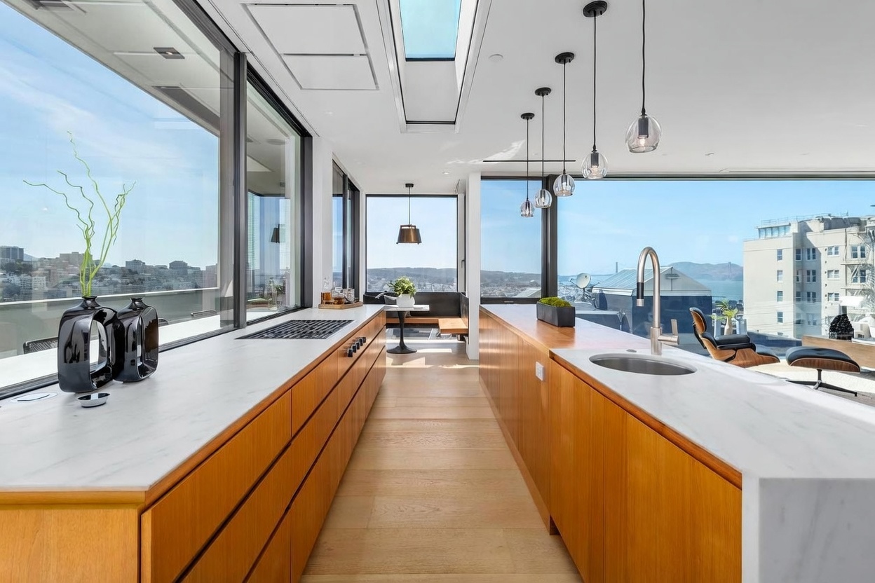 This sleek, modern kitchen features warm-toned wood cabinetry paired with light-colored stone countertops and a central island. The space is defined by expansive floor-to-ceiling windows that offer panoramic city and bay views, complemented by a skylight that floods the area with natural light. A series of minimalist pendant lights hangs above the island, creating a sophisticated atmosphere that seamlessly blends indoor luxury with the urban landscape.