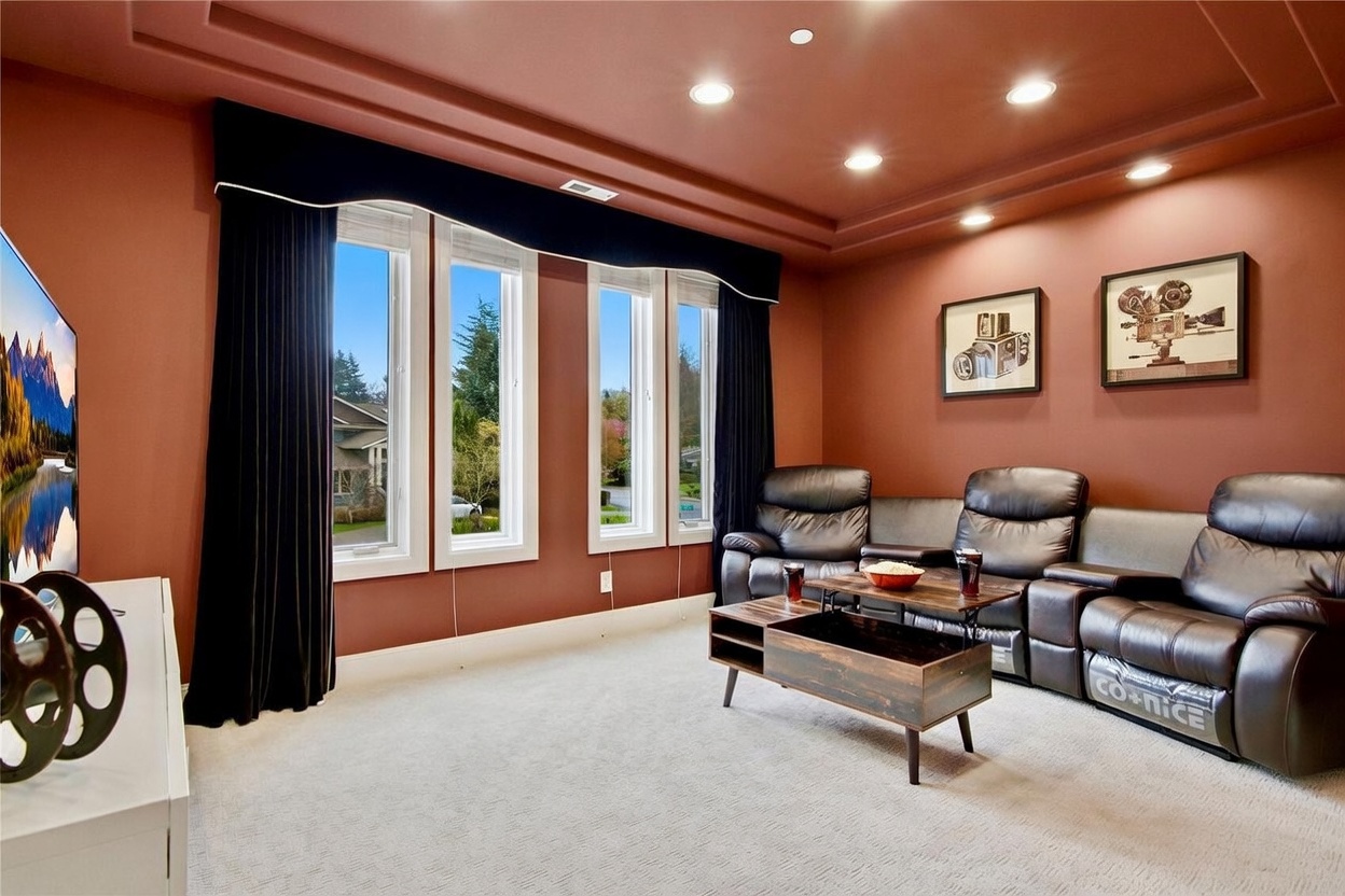 This media room features deep red walls, a coffered ceiling with recessed lighting, and plush dark leather theater-style seating. A central coffee table with a lift-top design sits on light-colored carpeting, while large windows with dark curtains provide natural light and a view of the neighborhood. The room is decorated with framed vintage cinema-themed artwork, creating a dedicated space for entertainment.