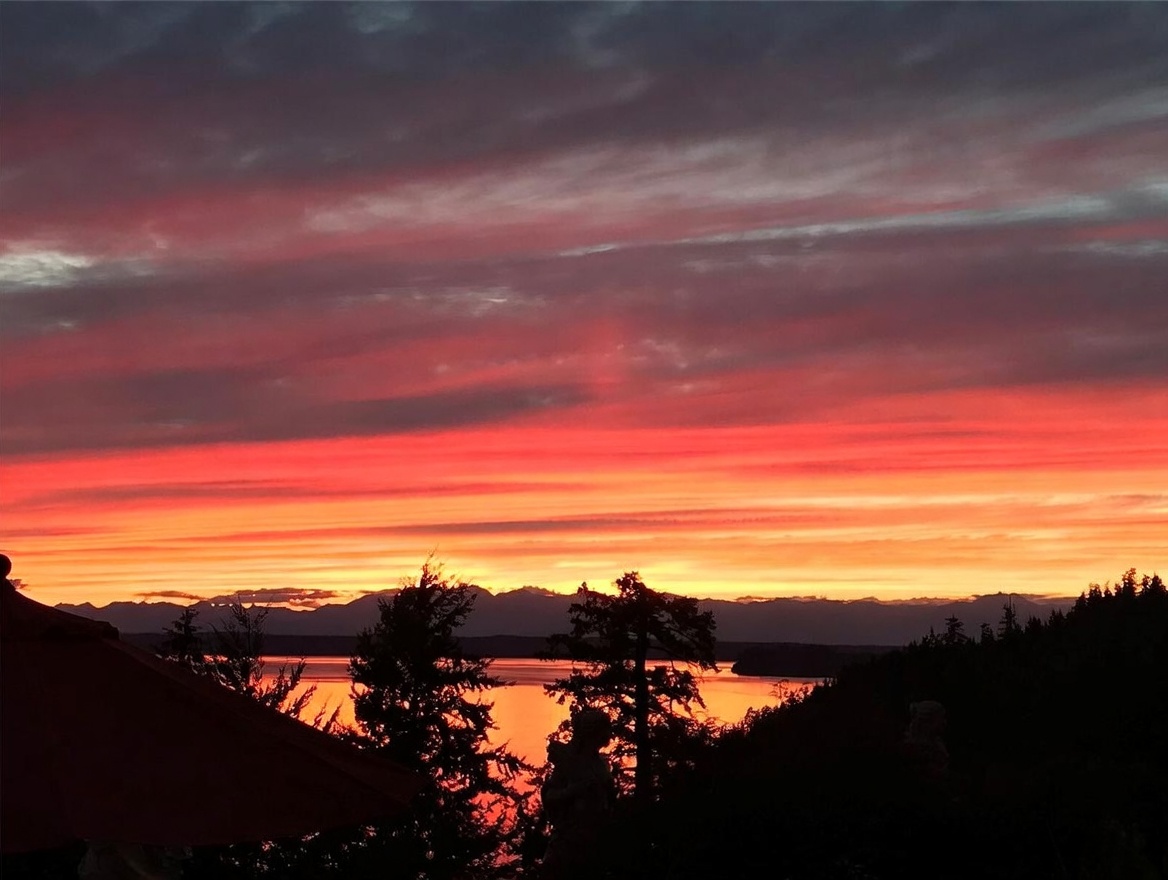 This image captures a breathtaking sunset view from an elevated vantage point, likely a deck or balcony. The horizon is dominated by a vibrant, multi-colored sky reflecting over a calm body of water, with silhouettes of distant mountains and foreground trees framing the scene. The perspective suggests a serene, high-end outdoor living space designed to maximize scenic vistas.