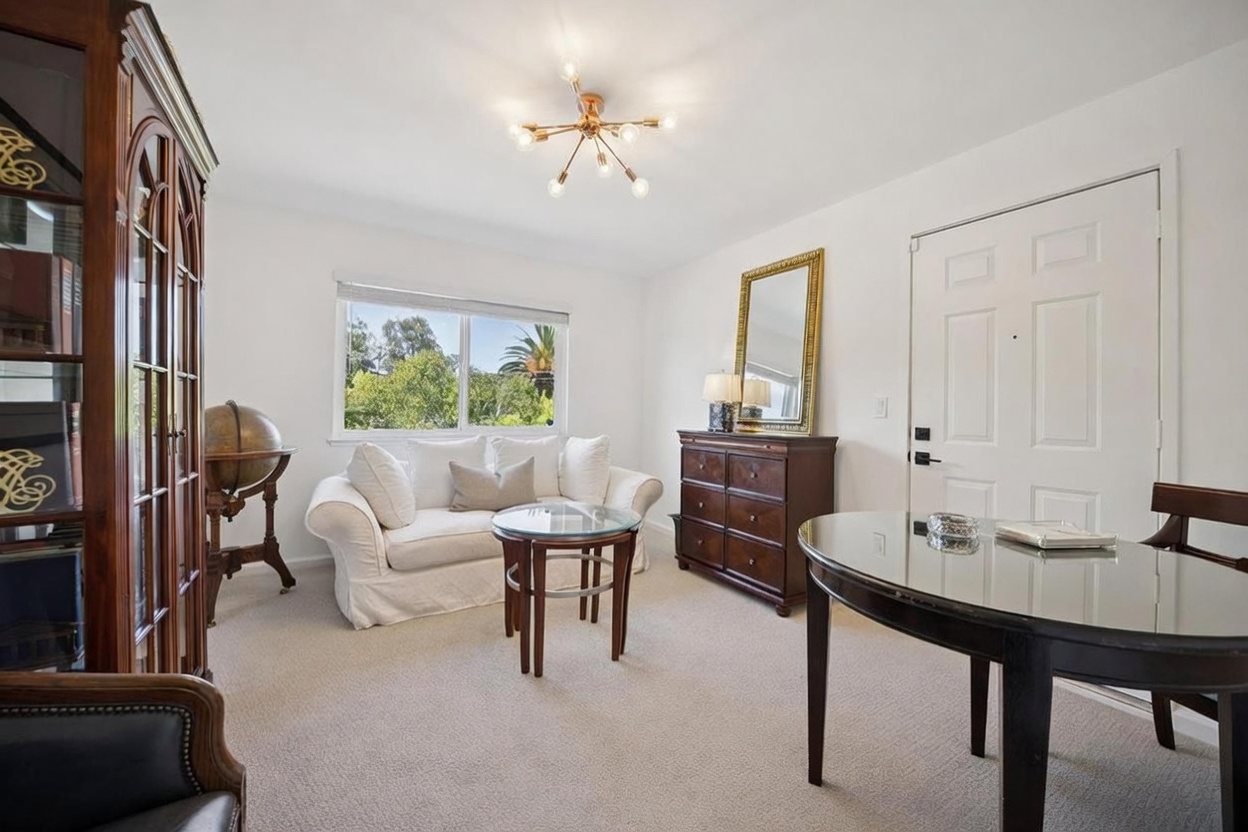 This bright and inviting room functions as a versatile study or sitting area, featuring a plush white sofa, a glass-topped coffee table, and a dark wood dresser topped with a large gold-framed mirror. A globe on a stand adds a classic touch, while a round glass-topped table in the foreground provides a functional workspace. The space is illuminated by a modern, multi-arm chandelier and benefits from natural light streaming through a large window overlooking greenery.