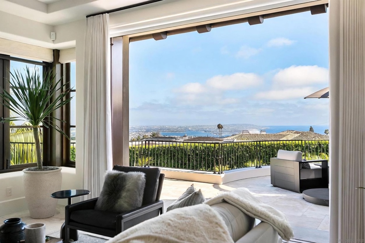 This elegant living space features a seamless transition to an expansive outdoor terrace through large, open glass doors that frame a panoramic coastal view. The interior is furnished with a comfortable dark armchair and a plush sofa, while a large potted plant adds a touch of greenery to the bright, airy room. The outdoor area includes a stone-paved balcony with a metal railing, overlooking a scenic landscape of hills and the ocean beyond.
