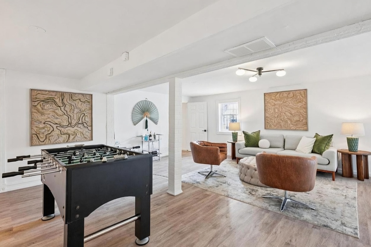 This spacious, open-concept basement features a versatile layout that combines a game area with a foosball table and a comfortable lounge space. The room is anchored by a light-toned wood-look floor, a neutral-colored sofa, and two leather swivel chairs arranged around a tufted ottoman. Decorative textured wall art and a unique ceiling light fixture add character to the bright, white-walled interior.