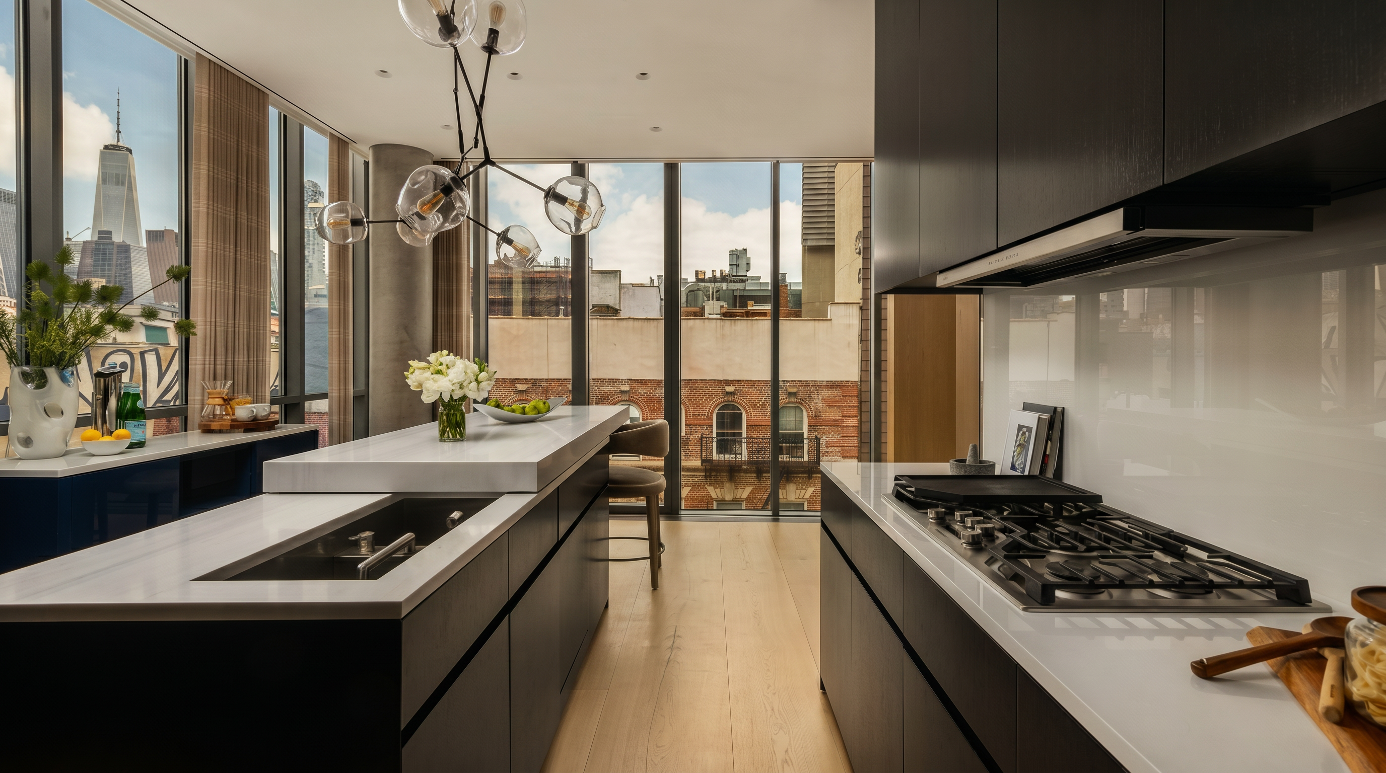 This modern, high-end kitchen features sleek dark cabinetry contrasted by light stone countertops and light wood flooring. A large central island with an integrated sink and seating area serves as the focal point, illuminated by a sculptural, multi-globe pendant light. The space is framed by expansive floor-to-ceiling windows that offer a striking urban view, including a prominent skyscraper, creating a bright and sophisticated atmosphere.