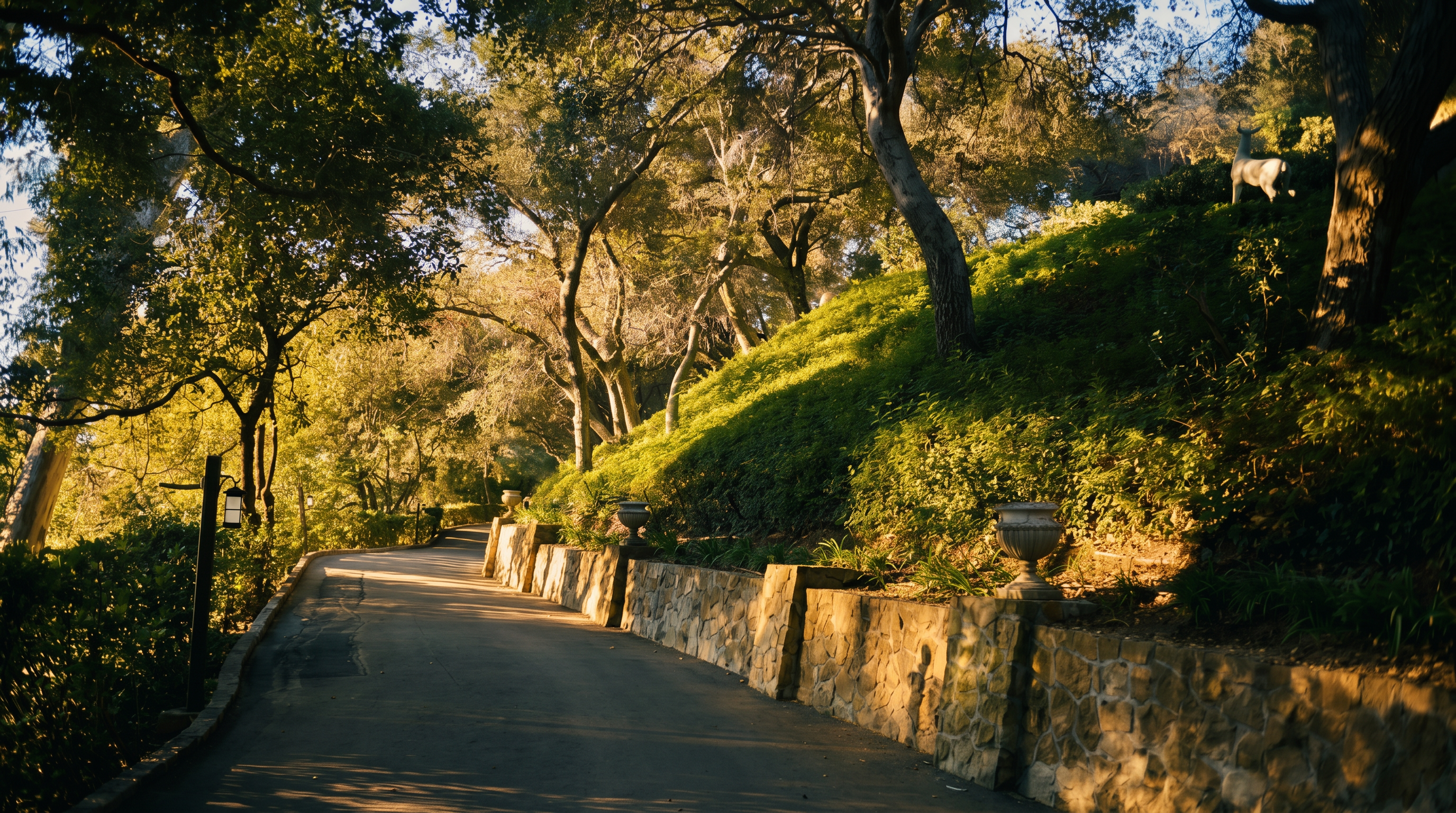 This scenic outdoor pathway winds through a lush, tree-lined landscape, featuring a stone retaining wall adorned with decorative urns. The perspective captures a serene, sun-dappled walkway that leads deeper into the property, highlighting the mature trees and manicured greenery. A small deer statue adds a whimsical touch to the hillside, creating an inviting and tranquil atmosphere.