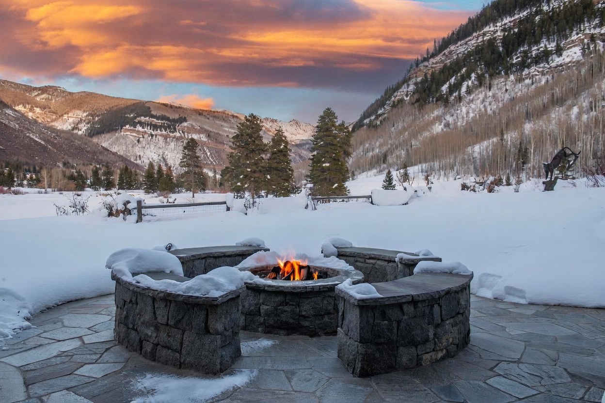 This image captures a serene outdoor patio featuring a circular stone fire pit surrounded by curved stone benches, all dusted with a light layer of snow. The foreground provides a cozy, intimate seating area that overlooks a vast, snow-covered landscape framed by towering pine trees and majestic, rugged mountains under a vibrant sunset sky. The perspective is low and centered, emphasizing the inviting warmth of the fire against the cold, expansive winter backdrop.