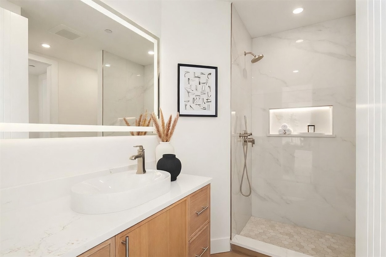 This modern bathroom features a sleek vanity with a light wood finish, a white vessel sink, and a large, illuminated mirror. To the right, a glass-enclosed walk-in shower showcases light-colored stone wall tiles and a built-in recessed niche with accent lighting. The space is decorated with minimalist art and a decorative vase, creating a clean, contemporary aesthetic.