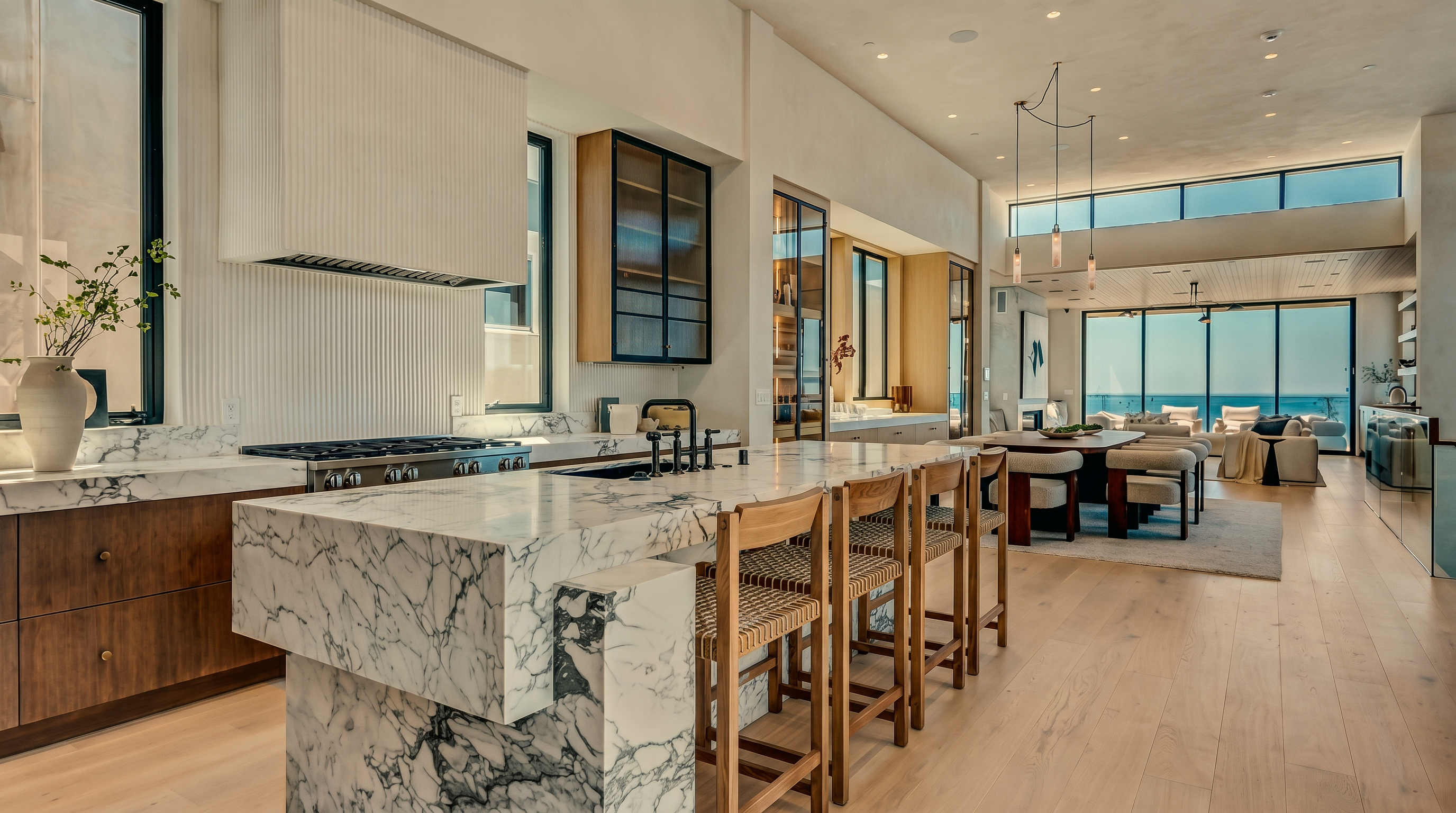 This expansive, open-concept kitchen features a large central island with a waterfall-edge stone countertop and integrated seating. The space is characterized by light wood flooring, minimalist cabinetry, and a prominent range hood, all bathed in natural light from large windows that overlook the ocean. The perspective captures the seamless flow from the culinary workspace into the adjacent dining and living areas, emphasizing a bright, airy, and sophisticated coastal aesthetic.
