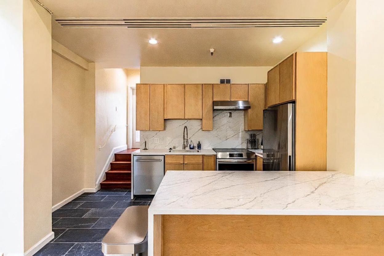This modern kitchen features light wood cabinetry, stainless steel appliances, and a stone-topped island that provides a functional workspace. A marble-patterned backsplash adds a sophisticated touch, while the dark tile flooring creates a striking contrast with the light-toned cabinets. The perspective looks from the island toward the kitchen area, with a set of wooden stairs visible in the background leading to another level of the home.