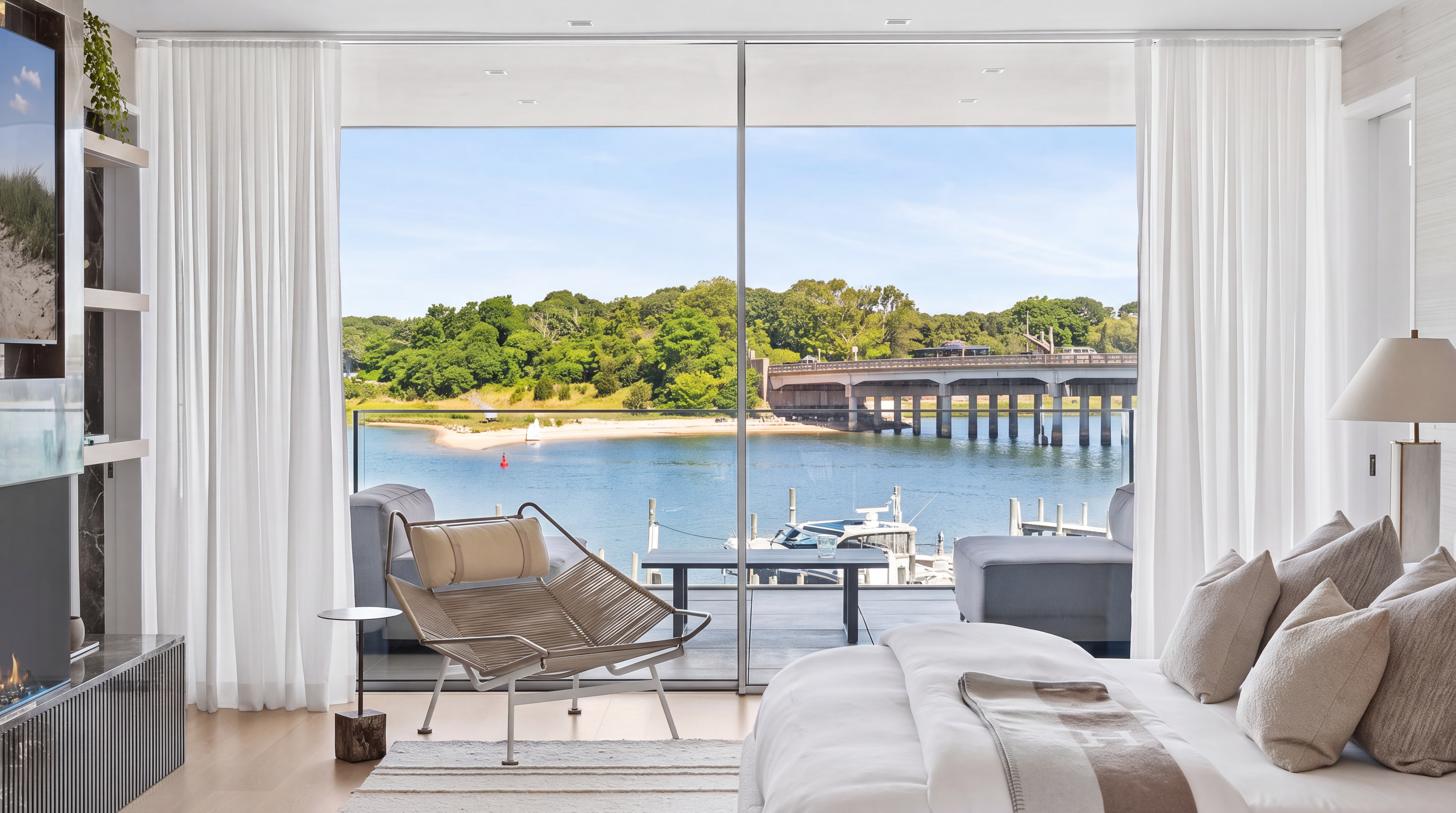 This modern, luxury primary bedroom features high-end finishes, including a minimalist fireplace, sleek shelving, and large floor-to-ceiling glass sliding doors. The room seamlessly flows out to a private balcony that offers breathtaking, panoramic views of the water, a bridge, and surrounding lush greenery. The interior aesthetic is sophisticated and serene, characterized by a neutral palette and clean lines that highlight the spectacular waterfront setting.