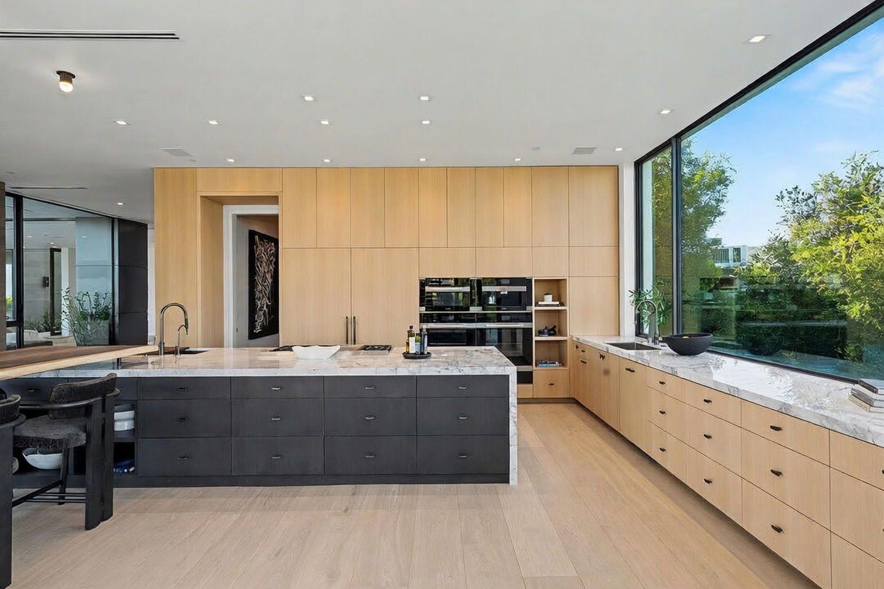 This modern, minimalist kitchen features a large central island with a dark base and a light stone countertop, complemented by extensive light wood cabinetry. A long run of matching light wood cabinets lines the wall, incorporating built-in appliances and a sink positioned beneath a expansive window that offers a clear view of the outdoors. The space is bright and airy, characterized by clean lines, recessed lighting, and a seamless integration of natural light.