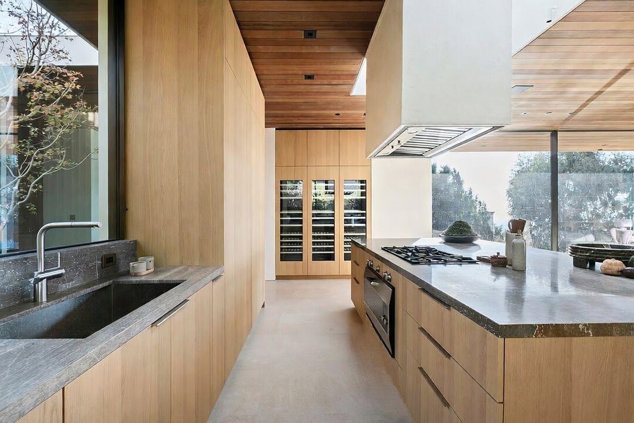 This sleek, modern kitchen features warm wood cabinetry, stone countertops, and a large central island with a built-in gas cooktop and oven. The space is defined by its clean lines, a dramatic wooden ceiling, and floor-to-ceiling glass walls that seamlessly blend the interior with the outdoor landscape. A dedicated wine storage area is visible in the background, contributing to the sophisticated and high-end aesthetic of the home.