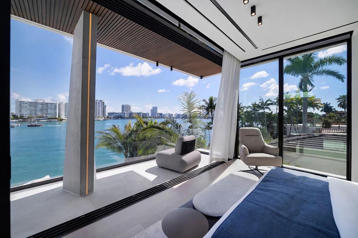This luxurious primary bedroom features expansive floor-to-ceiling glass sliding doors that open onto a private balcony overlooking a scenic waterfront cityscape. The interior is minimalist and modern, furnished with a plush armchair and a sleek bed, while the balcony includes a contemporary lounge chair. The seamless transition between the indoor space and the outdoor terrace creates an airy, sophisticated atmosphere with panoramic views.