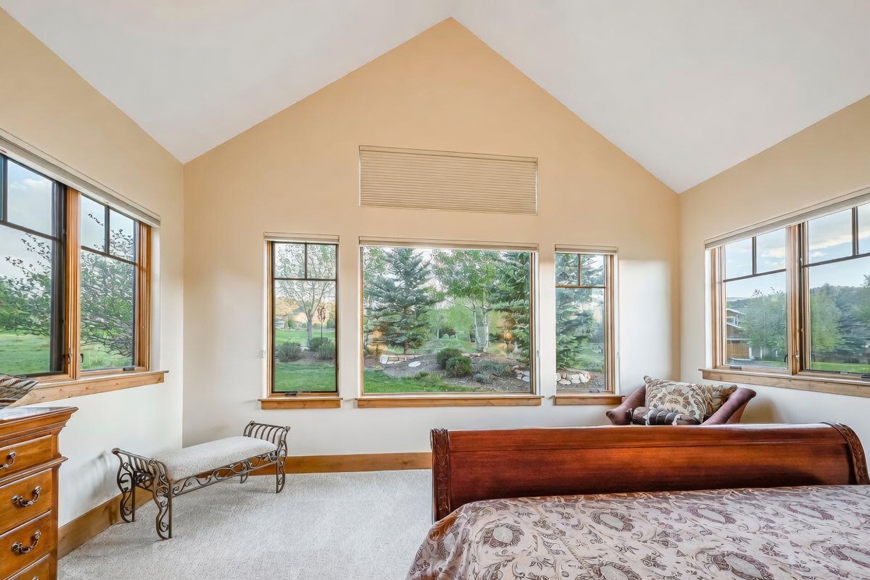 This bright and airy bedroom features a vaulted ceiling and a series of large windows that provide expansive views of the surrounding landscape. The room is furnished with a wooden bed frame and a small bench, creating a peaceful and inviting atmosphere. The neutral color palette and natural light enhance the sense of space and tranquility in this primary suite.