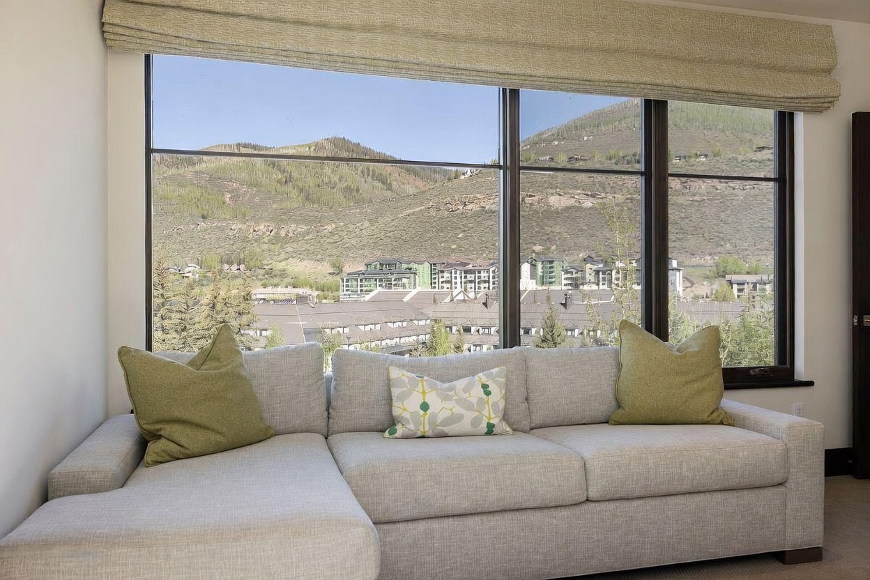 This living room features a large, light-colored sectional sofa positioned directly in front of expansive windows that frame a scenic mountain and town view. The room is accented with two solid green throw pillows and one patterned accent pillow, creating a comfortable and inviting seating area. The neutral color palette and clean lines emphasize the natural light and the impressive landscape visible through the glass.