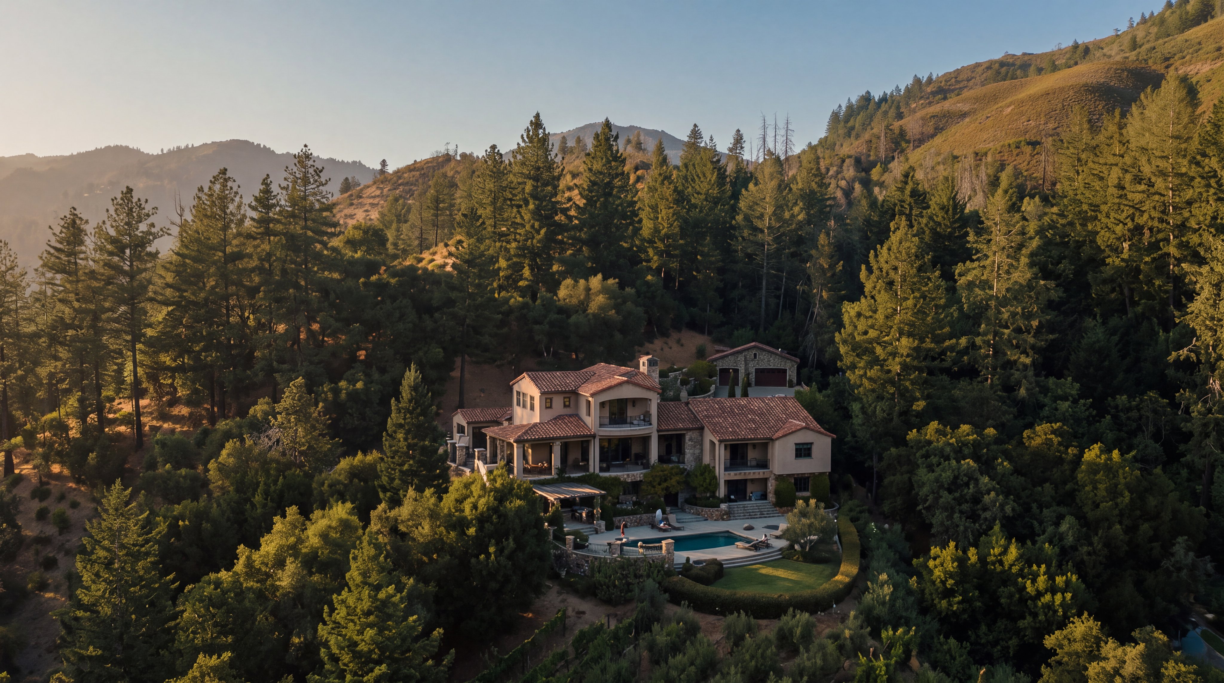 This high-angle aerial view captures a sprawling Mediterranean-style estate nestled into a lush, forested hillside. The multi-level residence features warm-toned stucco walls, a terracotta tile roof, and a private swimming pool set within a landscaped terrace. The property is surrounded by dense woodland and rolling hills, emphasizing its secluded and serene mountain setting.