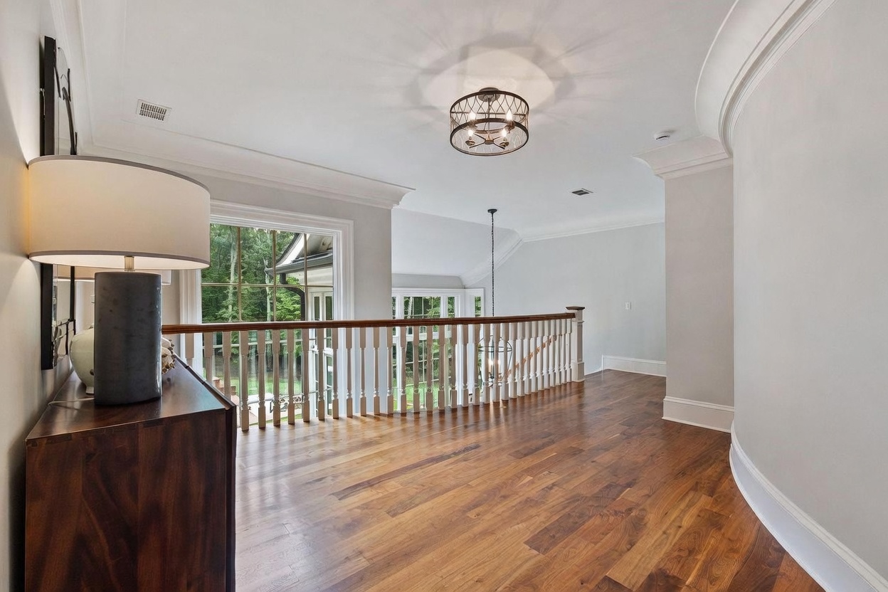 This bright and airy landing area features rich wood flooring and a classic white railing that overlooks the lower level. A curved wall adds architectural interest, while a decorative drum-style chandelier provides elegant overhead lighting. Large windows offer a view of the surrounding trees, creating a serene and open atmosphere.