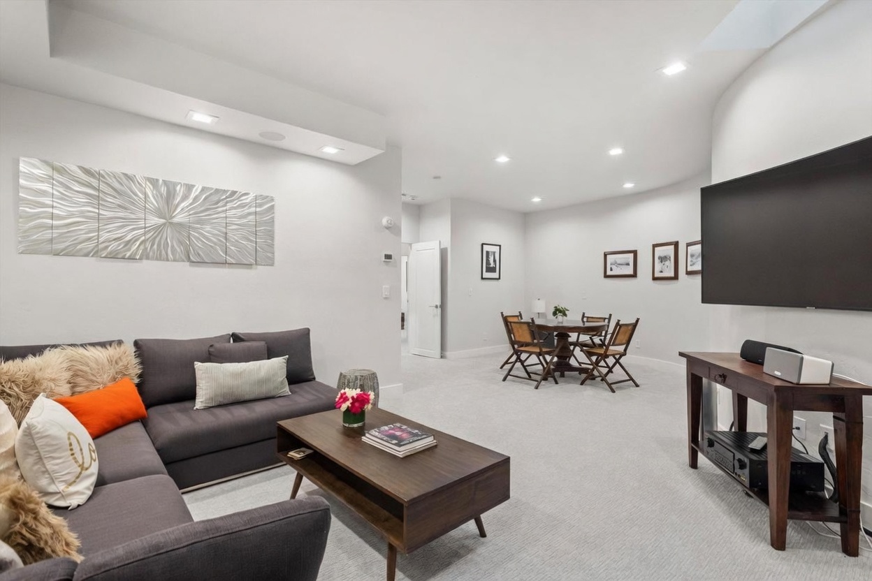 This open-concept living area features a dark gray sectional sofa accented with textured pillows and a central wooden coffee table. The space flows seamlessly into a dining area with a round table and four chairs, all set against a neutral color palette with recessed lighting. A large wall-mounted television sits above a wooden media console, creating a functional and inviting entertainment space.