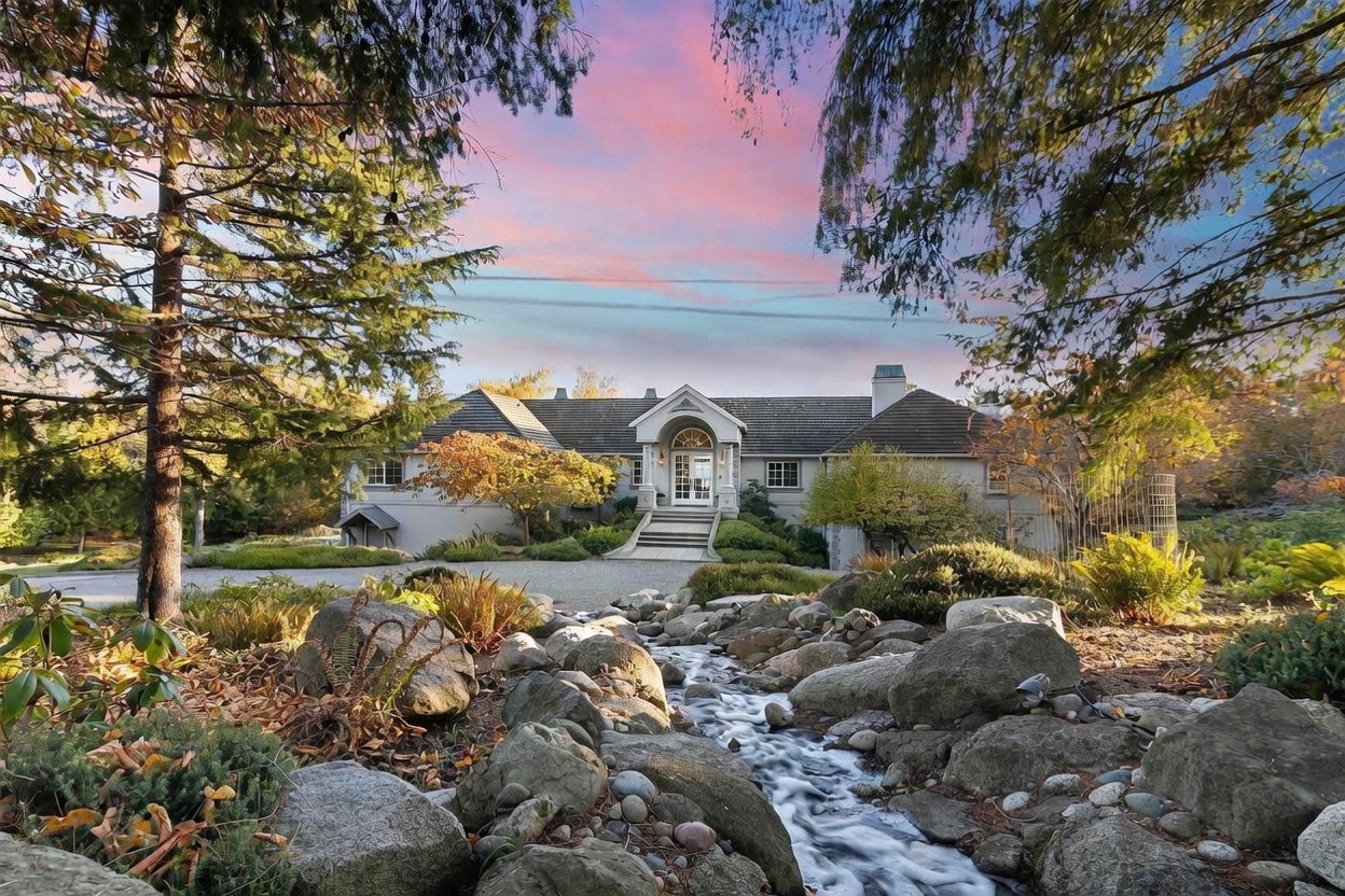 This elegant estate features a stately home with a light-colored facade and a prominent arched entryway, set against a backdrop of a vibrant sunset sky. In the foreground, a picturesque stone-lined stream flows through a lush, landscaped garden, creating a serene and inviting atmosphere. The property is framed by mature trees, emphasizing its private and tranquil setting.