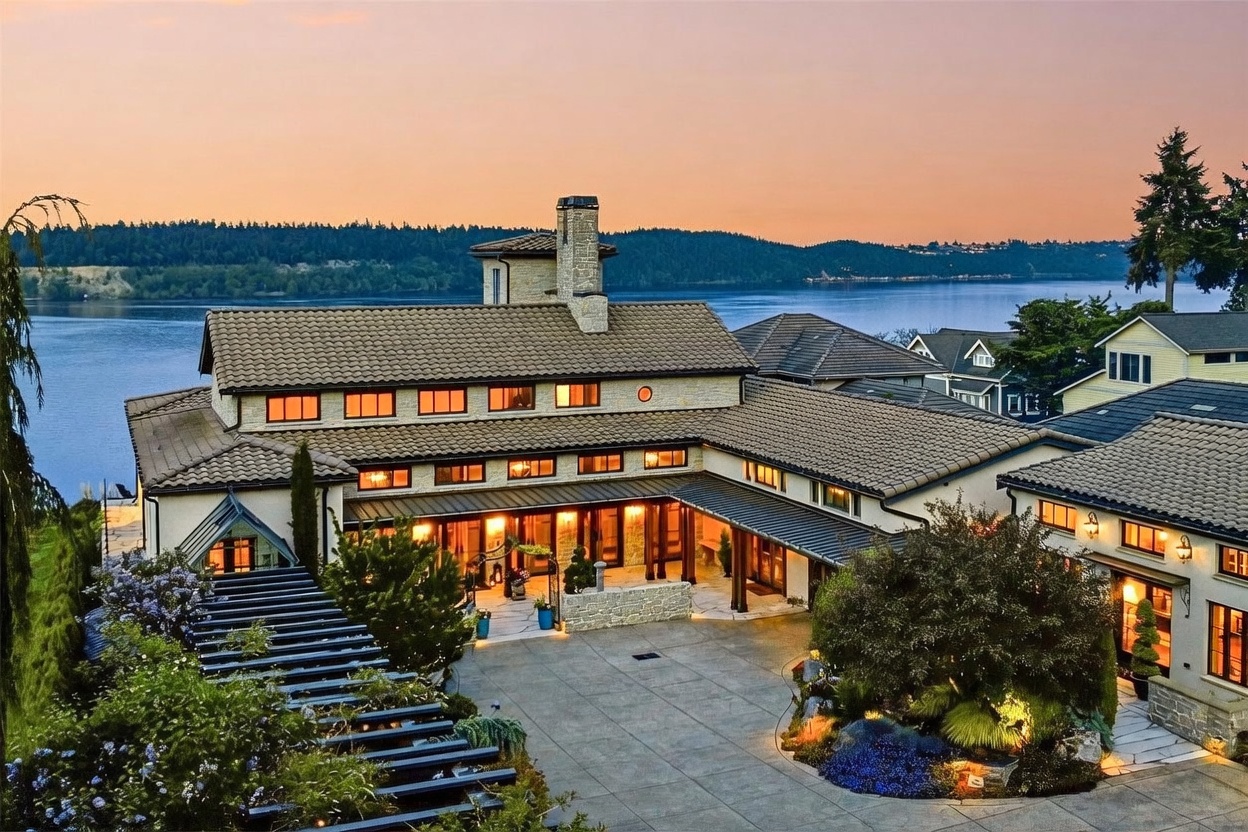 This elevated perspective captures a sprawling, multi-level estate at dusk, showcasing its Mediterranean-inspired architecture with a tiled roof and stone accents. The home features a large, paved motor court, a covered entryway, and a pergola-covered walkway, all set against a serene waterfront backdrop. The warm glow from the interior lighting contrasts beautifully with the soft twilight sky, emphasizing the property's luxurious and inviting atmosphere.