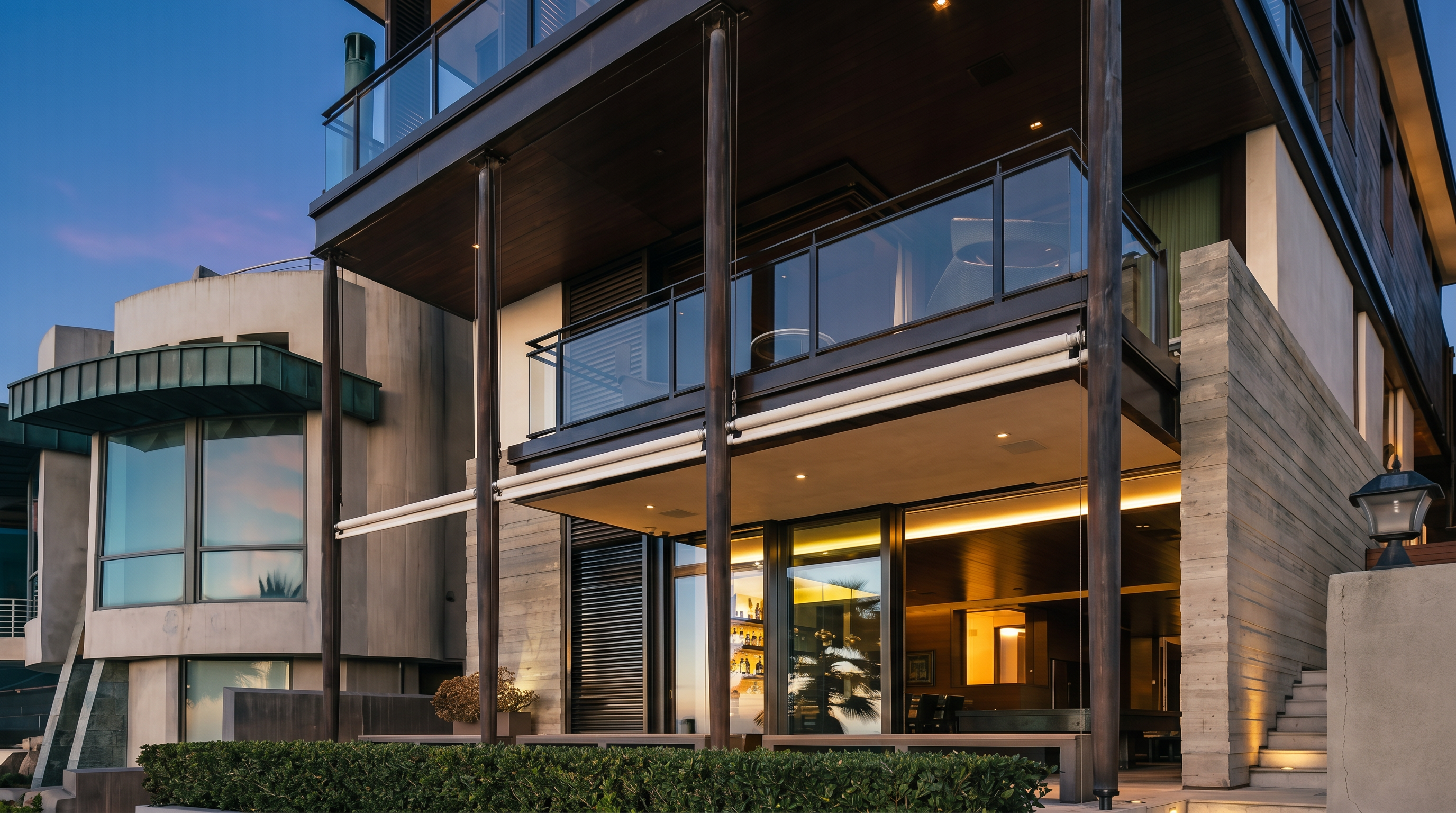 This modern, multi-story residence features a striking architectural design with dark metal framing, expansive glass balconies, and warm wood-toned accents. The ground level opens to a patio area, while the upper levels showcase floor-to-ceiling glass walls that emphasize an indoor-outdoor lifestyle. The structure is set against a clear twilight sky, highlighting the warm interior lighting that creates an inviting and sophisticated atmosphere.