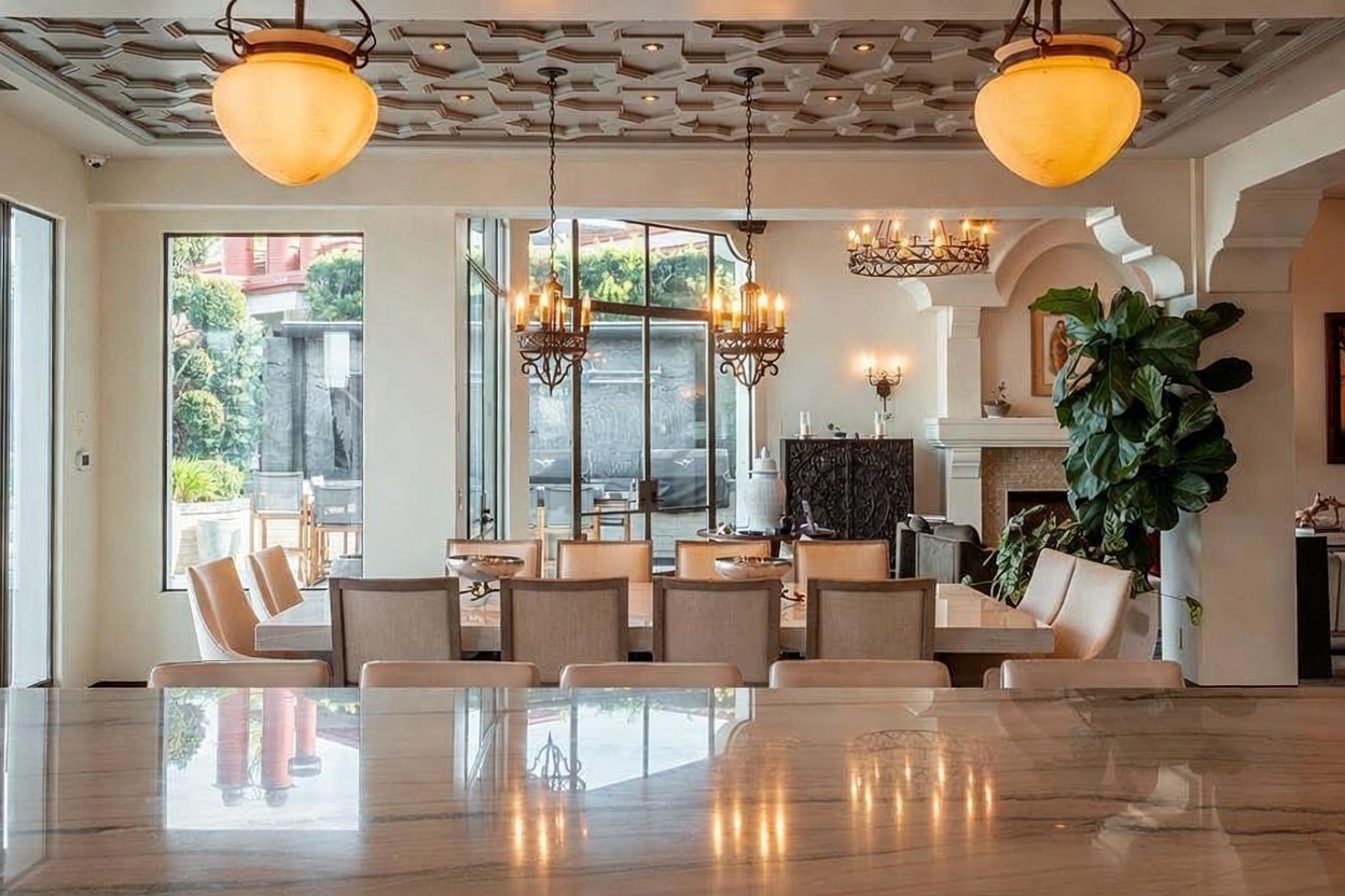 This elegant dining room features a long table surrounded by upholstered chairs, positioned beneath two large, warm-toned pendant lights. The space is framed by a coffered ceiling with intricate detailing and large windows that provide a view of the outdoor patio area. In the background, a fireplace with a decorative surround and a large potted plant add warmth and character to the sophisticated, open-concept layout.