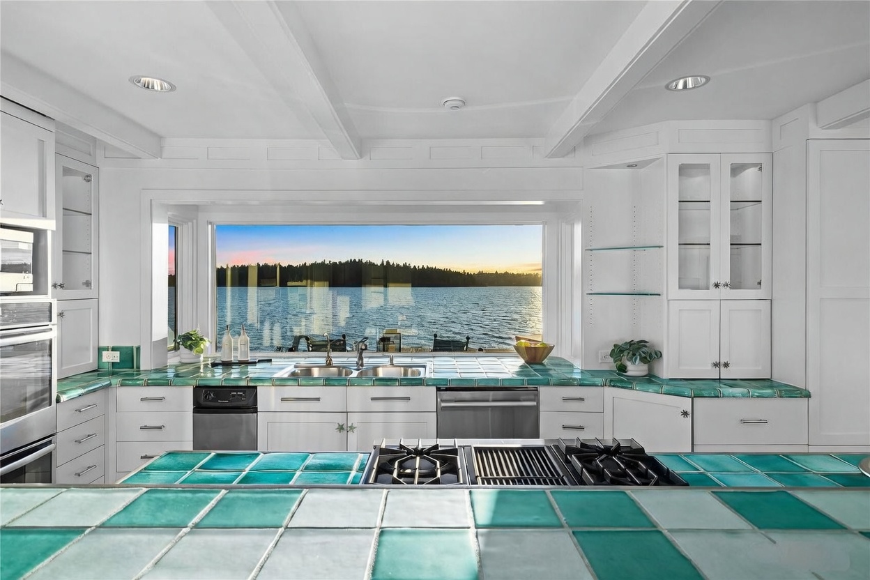 This bright, airy kitchen features a striking teal tiled countertop that contrasts beautifully with crisp white cabinetry and a large, panoramic window overlooking a serene lake. The perspective is centered from the kitchen island, showcasing a professional-grade cooktop and a seamless transition between the indoor workspace and the natural outdoor scenery. The overall aesthetic is a blend of classic coastal charm and functional design, perfect for enjoying waterfront views while cooking.