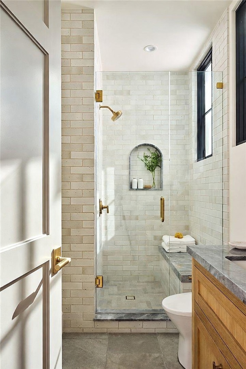 This elegant bathroom features a walk-in shower enclosed by a glass panel, showcasing light-colored brick-style wall tiles and a built-in stone bench. Brass fixtures, including the shower head and door handle, provide a warm contrast against the neutral palette, while an arched wall niche adds a decorative touch. The perspective from the doorway highlights the clean, modern design and the seamless transition between the vanity area and the shower space.