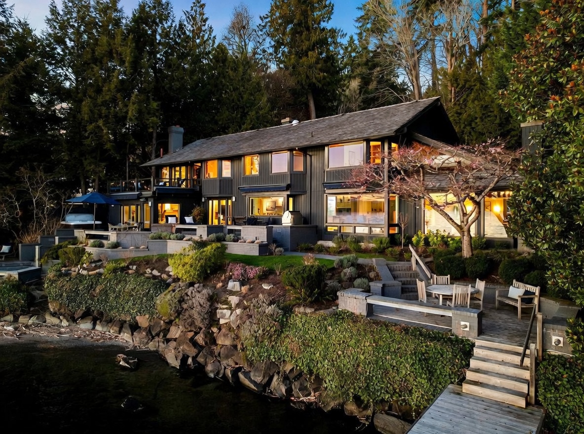 This stunning waterfront property features a multi-level dark-toned home nestled against a lush, forested hillside. The exterior showcases expansive windows, a tiered stone patio with integrated landscaping, and a private wooden staircase leading directly to the water's edge. The scene is captured at dusk, highlighting the warm, inviting glow from the interior lights against the natural, serene landscape.