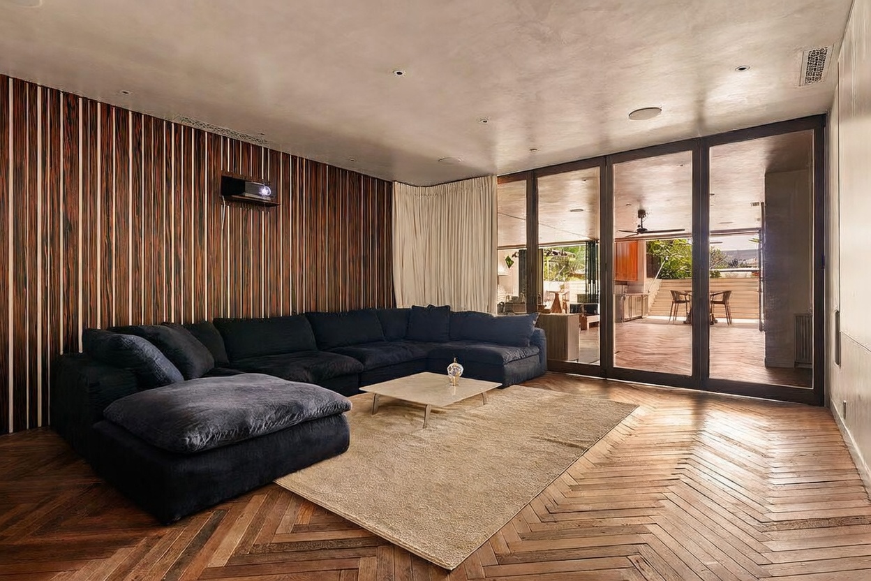 This sophisticated living room features a large, plush dark sectional sofa centered on a textured area rug atop rich, herringbone-patterned wood flooring. A striking wall with vertical wood paneling provides a warm, textured backdrop, while floor-to-ceiling glass doors open to an expansive outdoor patio area. The space is illuminated by recessed lighting and offers a seamless transition between indoor comfort and outdoor living.