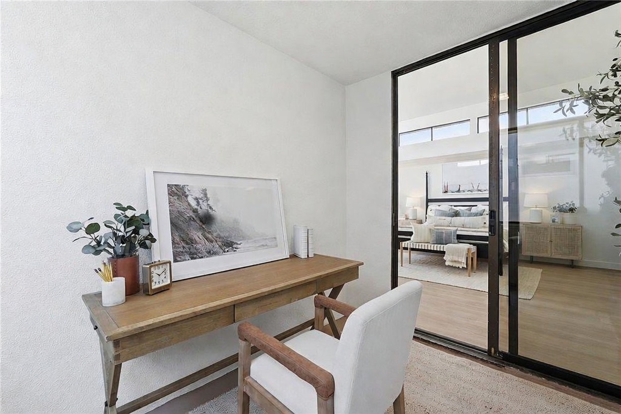 This bright home office space features a light wood desk paired with a modern white upholstered chair, creating a clean and minimalist aesthetic. A large framed landscape print sits on the desk, while a glass sliding door provides a clear view into an adjacent bedroom. The neutral color palette and natural light contribute to a calm, productive atmosphere.