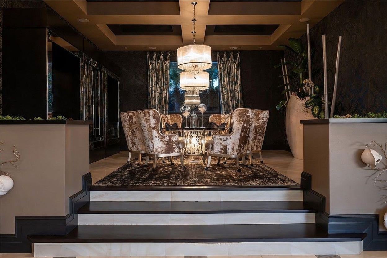 This formal dining room is elevated on a platform, accessed by two steps, and features a dramatic, dark-toned aesthetic. A central glass-topped table is surrounded by six plush, patterned chairs, all illuminated by a tiered crystal chandelier hanging from a coffered ceiling. The space is framed by dark walls with intricate wallpaper patterns, heavy curtains, and a large decorative planter, creating a sophisticated and moody atmosphere.