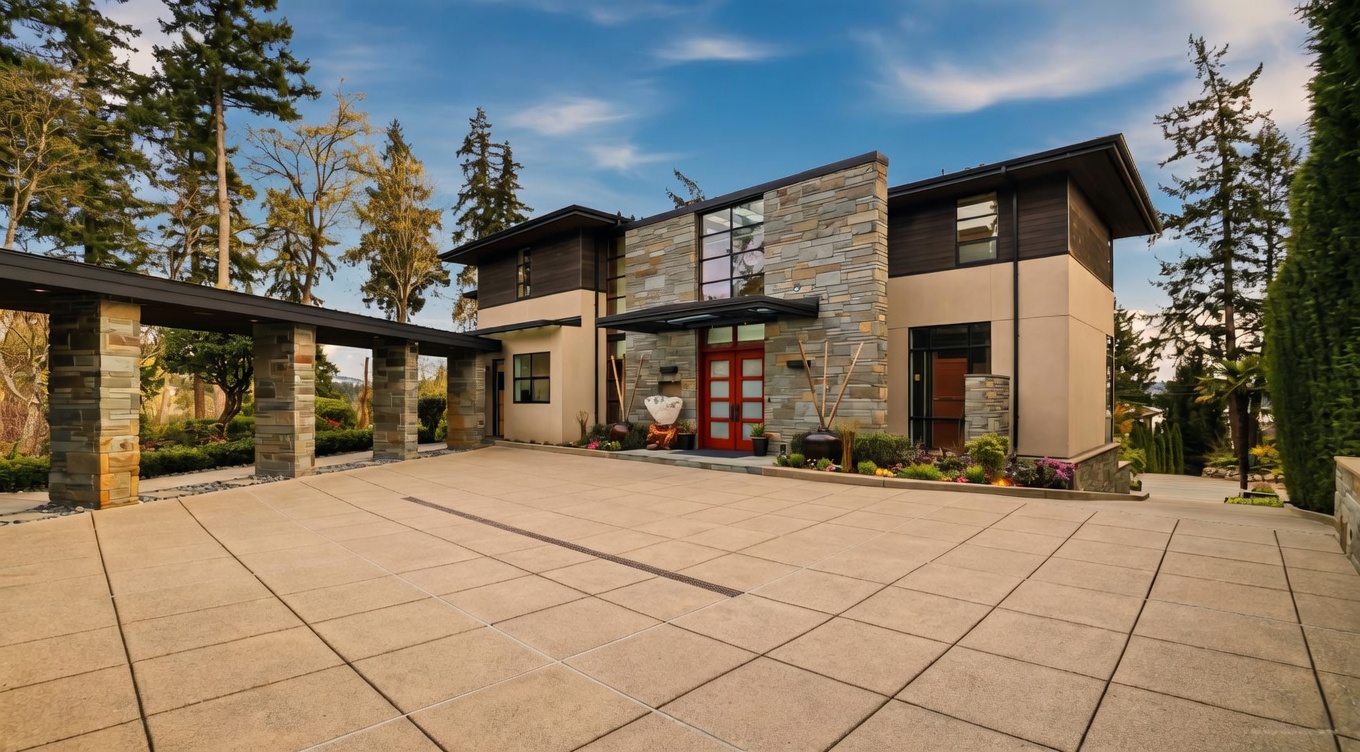 This modern residence features a striking facade characterized by a combination of stacked stone, smooth stucco, and dark wood accents. A prominent stone-clad entryway is framed by a covered walkway, leading to a vibrant red double-door entrance. The expansive concrete driveway provides a clean, geometric foreground, while the surrounding mature trees and clear blue sky create a serene and upscale atmosphere.