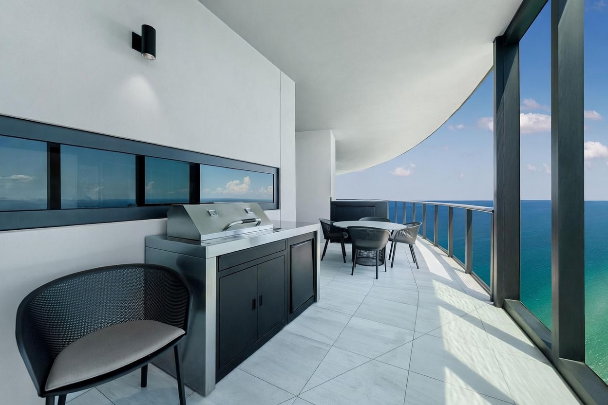 This sleek, modern balcony offers a panoramic view of the ocean through expansive glass railings. The space is equipped with a built-in stainless steel grill and a dining set, making it an ideal area for outdoor entertaining. The clean lines, neutral tile flooring, and minimalist aesthetic create a sophisticated and airy atmosphere.