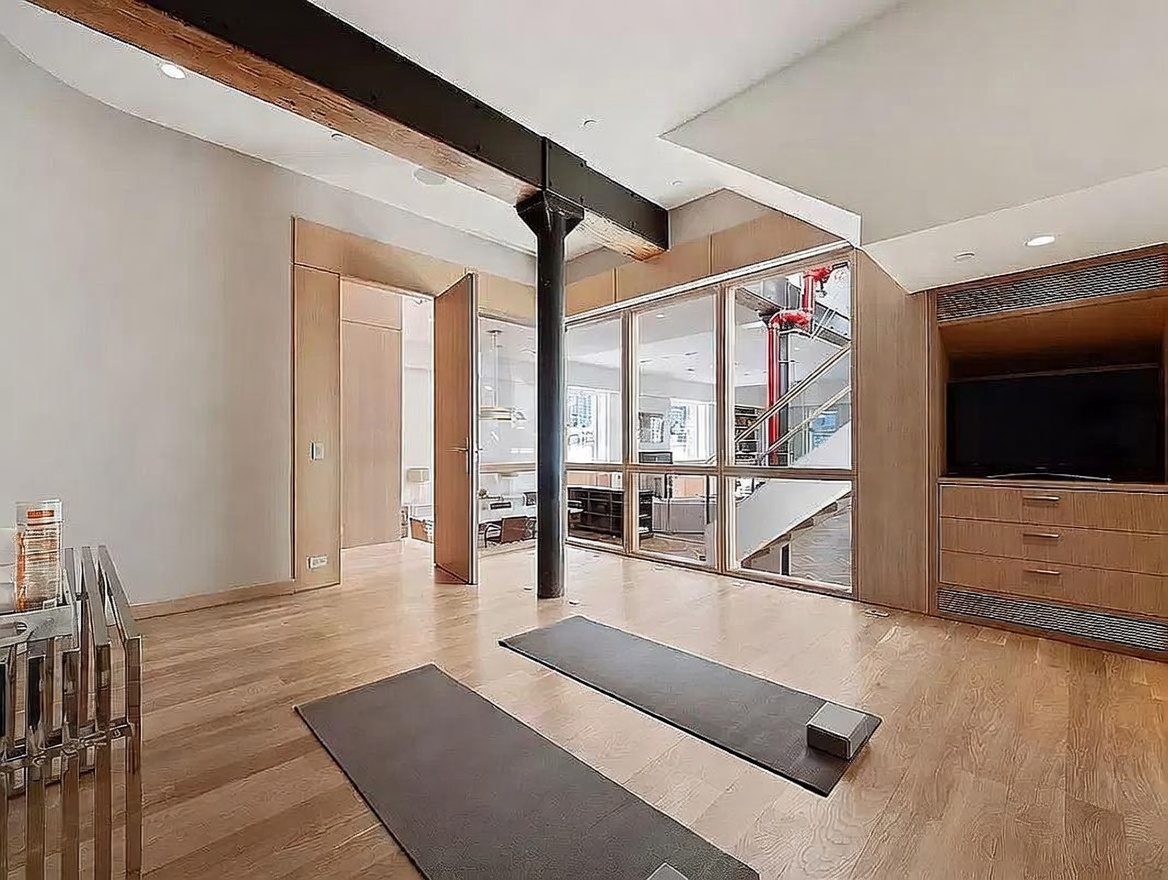 This bright, modern space is currently set up as a home gym, featuring two yoga mats laid out on light wood flooring. A prominent black structural column stands in the center of the room, while a glass-paneled wall provides a view into an adjacent living area with a staircase. To the right, a built-in light wood cabinetry unit houses a television, creating a clean and minimalist aesthetic.