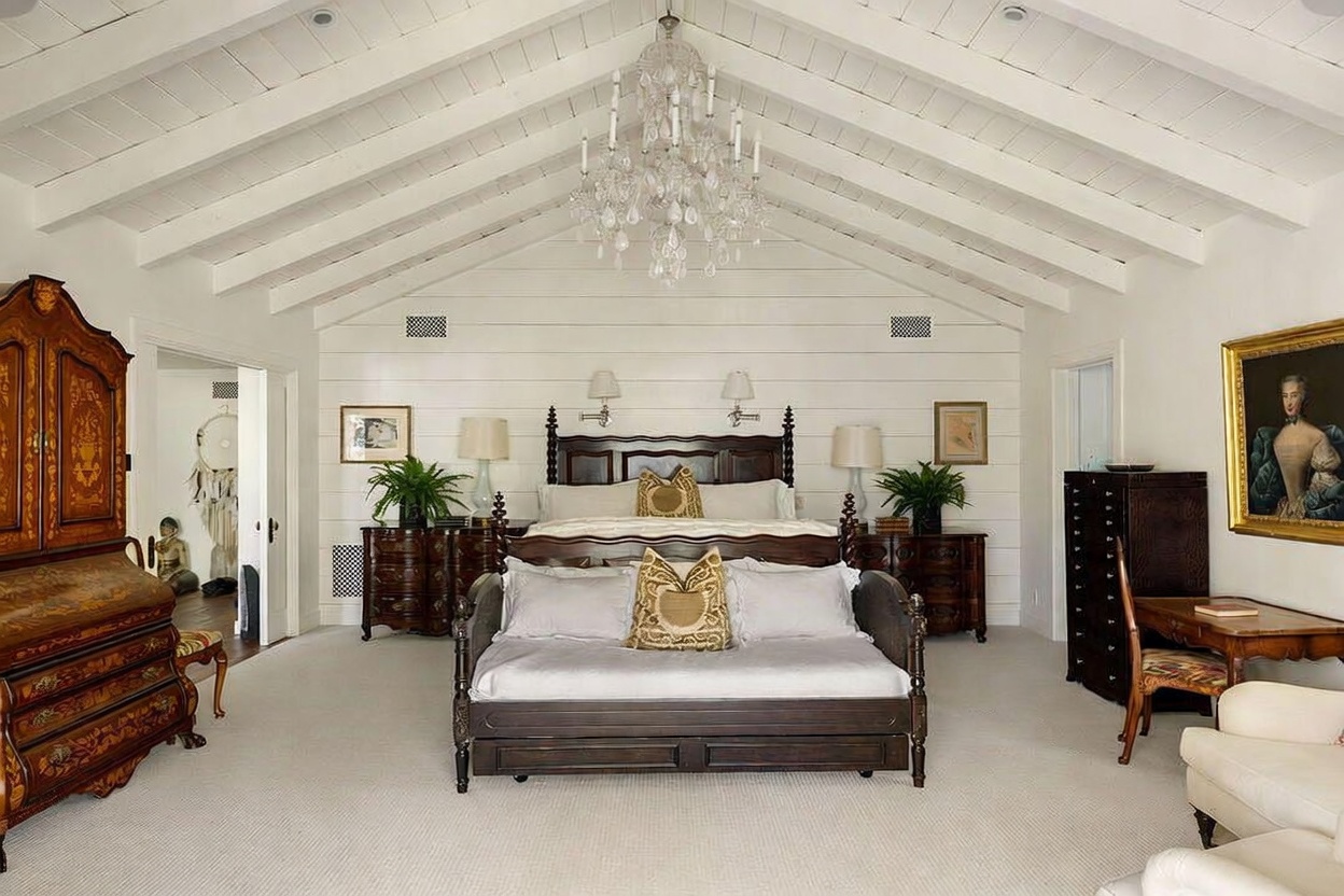 This spacious primary bedroom features a dramatic vaulted ceiling with exposed white beams and a large crystal chandelier as the central focal point. The room is anchored by a dark wood bed frame with a matching daybed at the foot, set against a white shiplap wall. Antique-style furniture, including a large armoire and a desk, adds a traditional touch to the bright, airy space.
