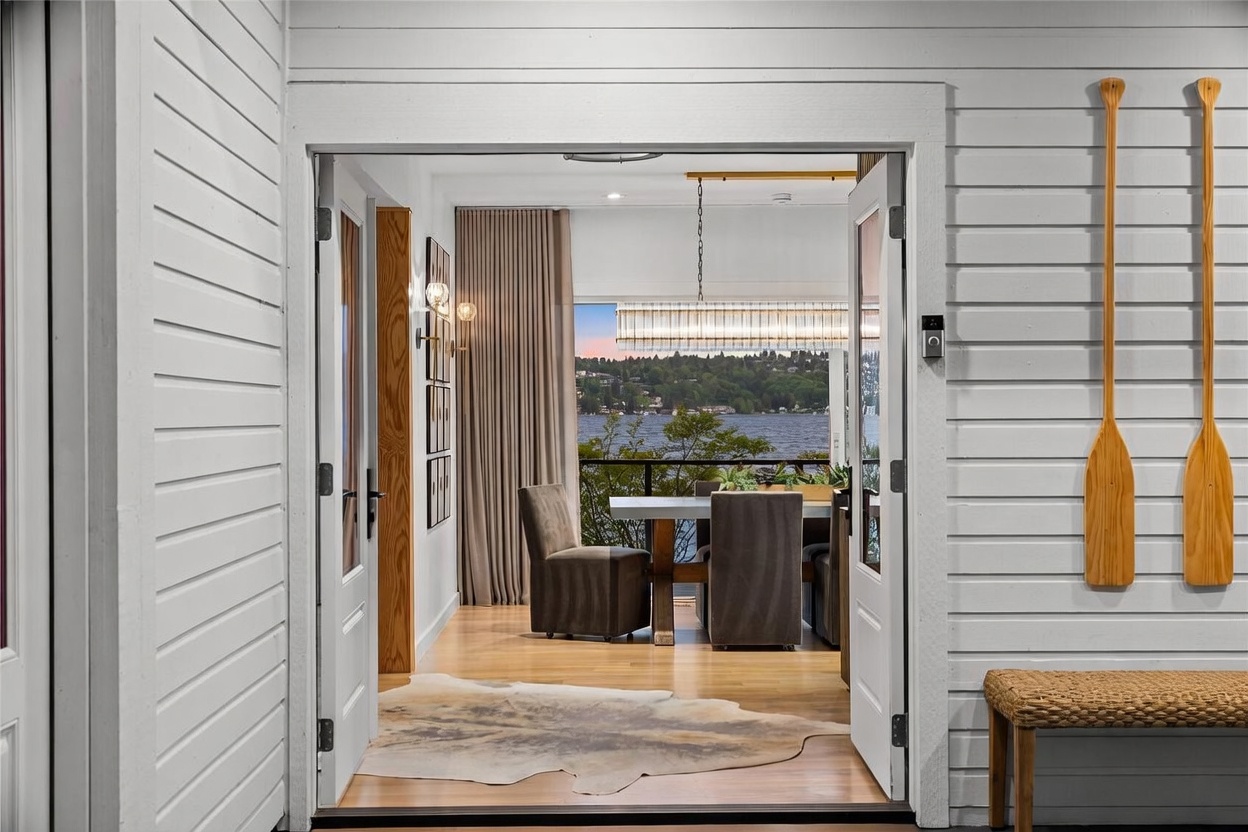 This perspective captures a welcoming entryway looking through open double doors into a bright dining area with a stunning lake view. The exterior features white horizontal siding and decorative wooden oars mounted on the wall, while the interior showcases light wood flooring, a cowhide rug, and a modern linear chandelier. The seamless transition from the outdoor porch to the indoor living space creates an inviting and airy atmosphere.