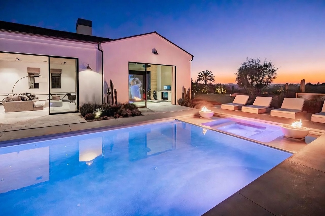 This stunning rear exterior view captures a modern luxury home at twilight, featuring a pristine swimming pool with integrated spa and two glowing fire bowls. Large sliding glass doors open from the interior living space and a dedicated golf simulator room, creating a seamless indoor-outdoor flow. The scene is framed by a sunset sky, desert-inspired landscaping, and a row of sleek lounge chairs, evoking a high-end resort atmosphere.