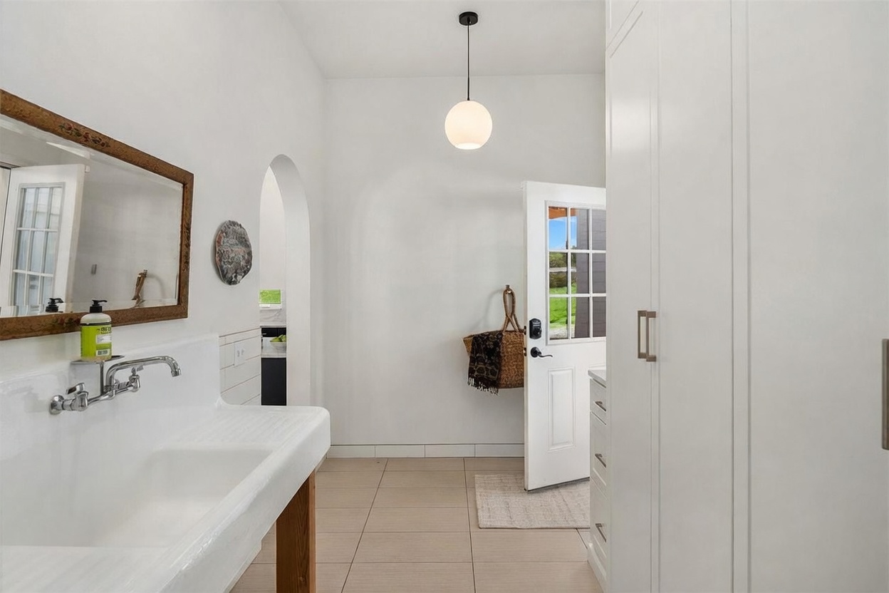 This bright, minimalist bathroom features a large, wall-mounted white utility-style sink with exposed plumbing fixtures. A tall, white storage cabinet stands to the right, while a framed mirror hangs above the sink, reflecting the clean, neutral aesthetic. An open doorway leads to an adjacent space, and a glass-paned door provides direct access to the outdoors, creating an airy and functional transition area.