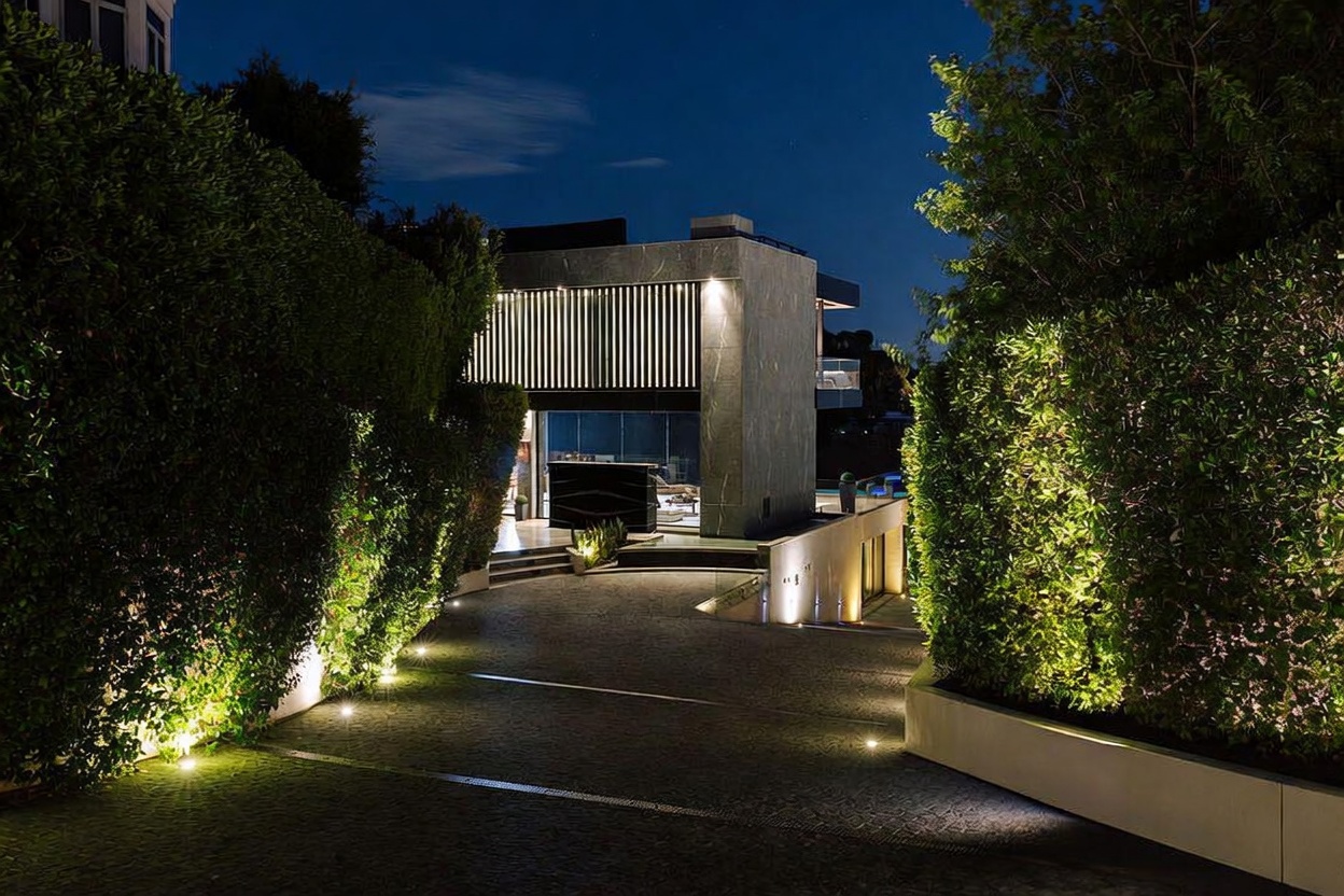 This striking modern residence is captured at twilight, showcasing a dramatic driveway approach flanked by lush, illuminated hedges. The architectural design features clean, geometric lines and a sophisticated stone facade, creating a luxurious and private atmosphere. The perspective emphasizes the grand entrance, drawing the viewer toward the glowing, contemporary structure.