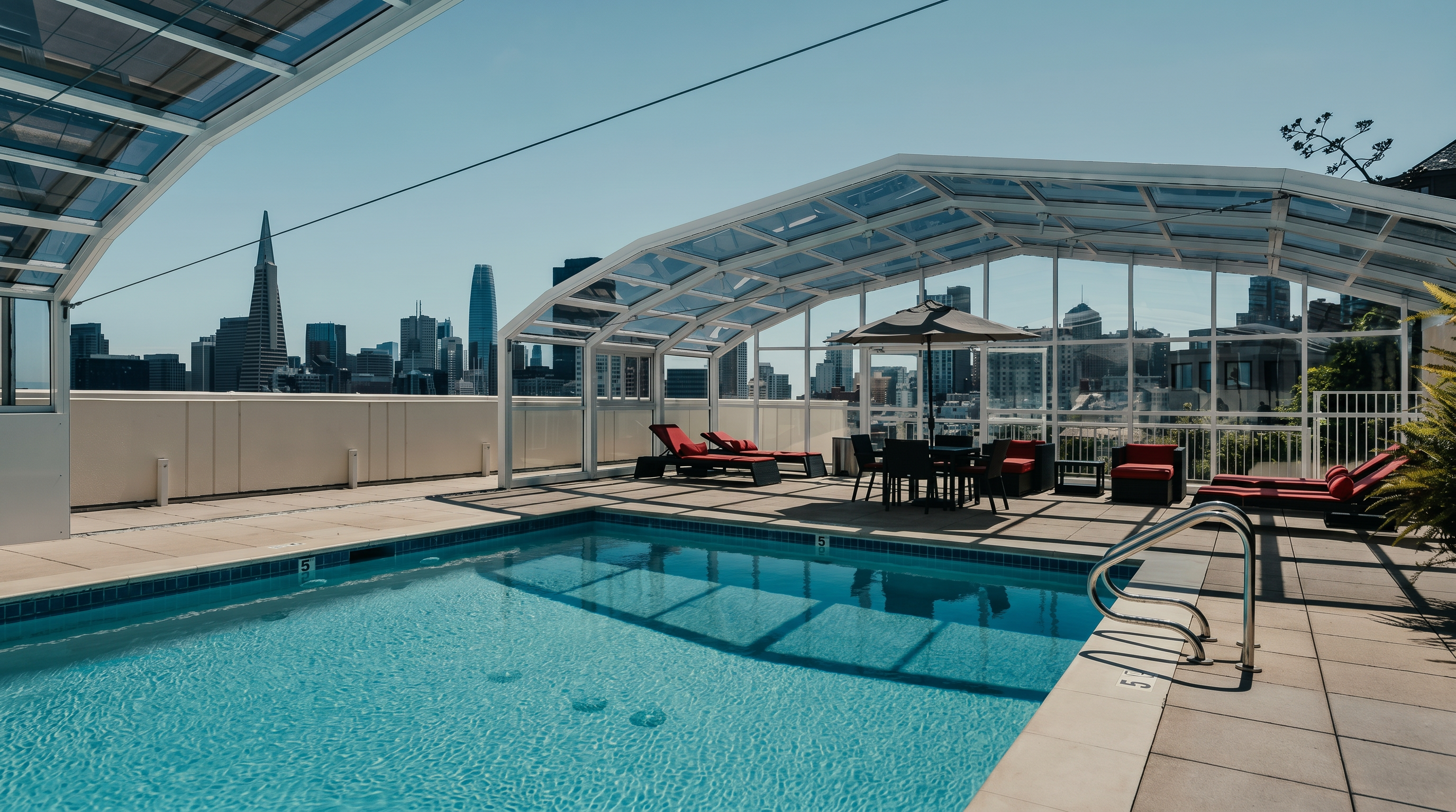 This rooftop pool area offers a stunning panoramic view of the San Francisco skyline, including the iconic Transamerica Pyramid. The space is partially enclosed by a retractable glass structure, providing a versatile environment for swimming and lounging. The deck is furnished with several red-cushioned lounge chairs and a dining set, creating an inviting outdoor retreat.