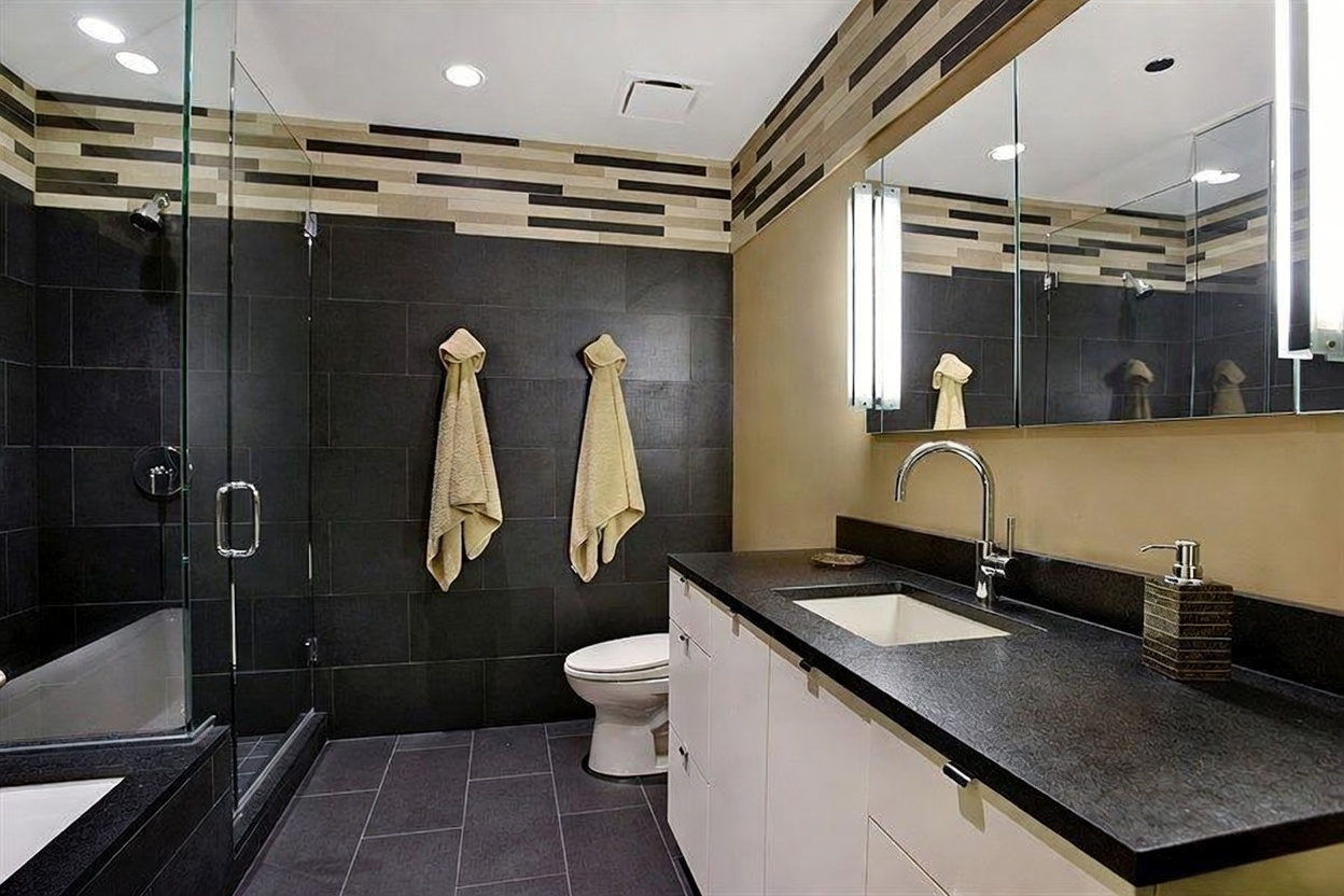 This modern bathroom features a sleek, dark-tiled walk-in shower with a glass enclosure and a contrasting vanity with a dark stone countertop. The walls are accented with a horizontal, multi-toned decorative tile border that adds texture to the space. A large mirror above the vanity enhances the room's brightness, while the minimalist white cabinetry provides a clean, contemporary aesthetic.