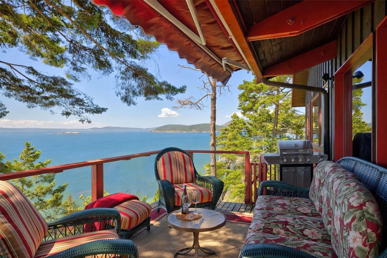 This elevated deck offers a stunning, panoramic view of the ocean and distant islands, framed by lush pine trees. The space is furnished with comfortable wicker seating featuring striped and floral cushions, centered around a small bistro table. A stainless steel grill is positioned to the side, making this an ideal spot for outdoor dining and relaxation while enjoying the coastal scenery.