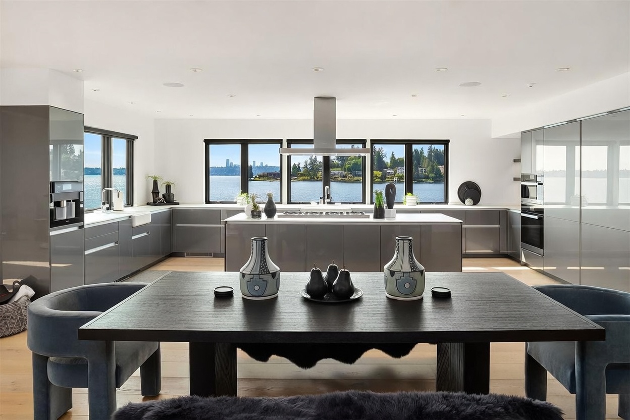This modern, open-concept kitchen features sleek, high-gloss grey cabinetry and a central island with a built-in cooktop. A large dining table with upholstered chairs sits in the foreground, while expansive windows behind the island provide a stunning, unobstructed view of the water and distant city skyline. The space is bright and airy, characterized by a minimalist aesthetic and high-end finishes.