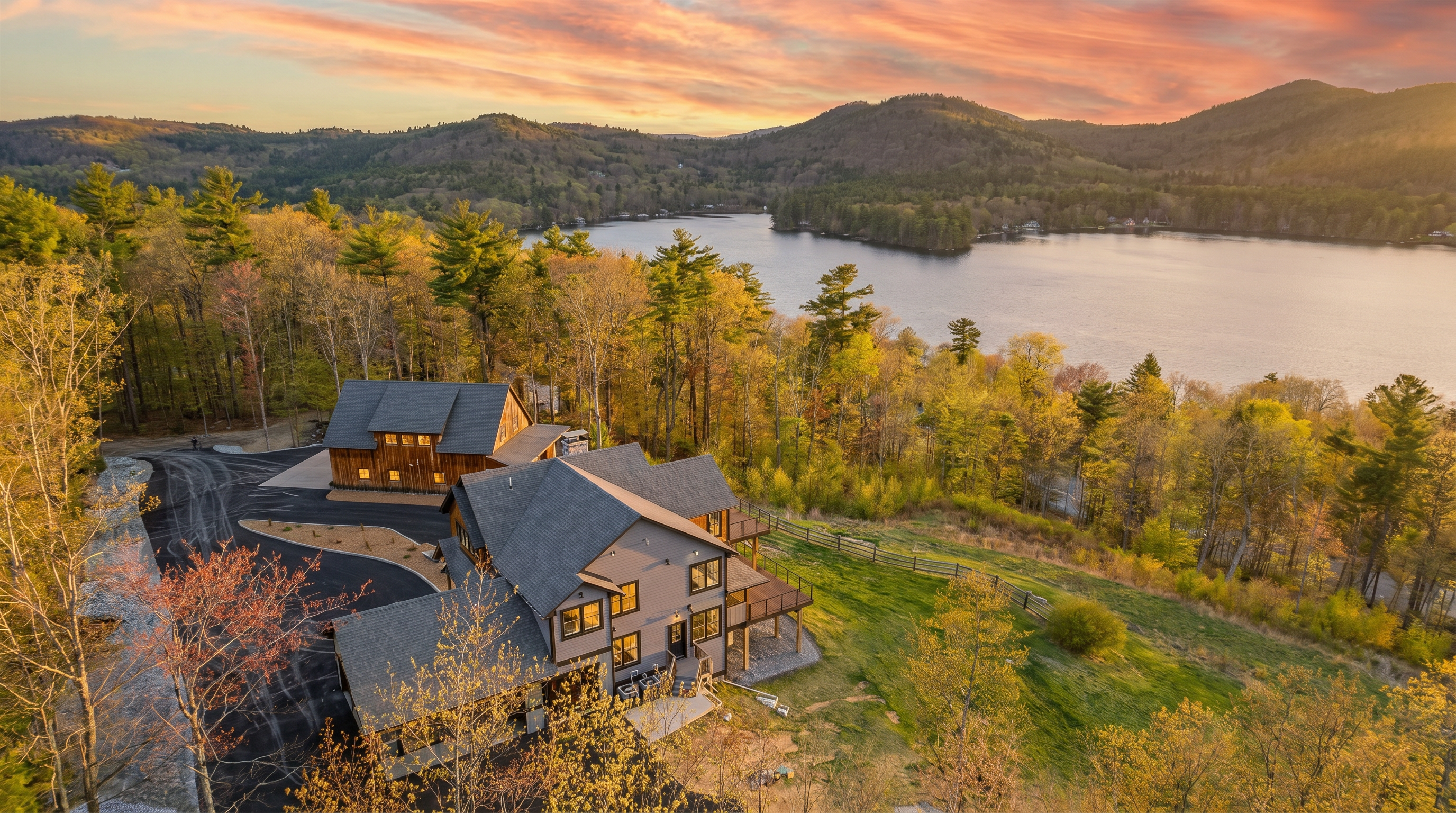 This high-angle aerial shot captures a multi-level gray-sided home nestled in a lush, wooded landscape overlooking a serene lake. The property features a paved driveway connecting the main residence to a separate wooden barn-style structure, with expansive green lawns and mature trees surrounding the site. The perspective emphasizes the home's integration into the natural environment and its proximity to the water.