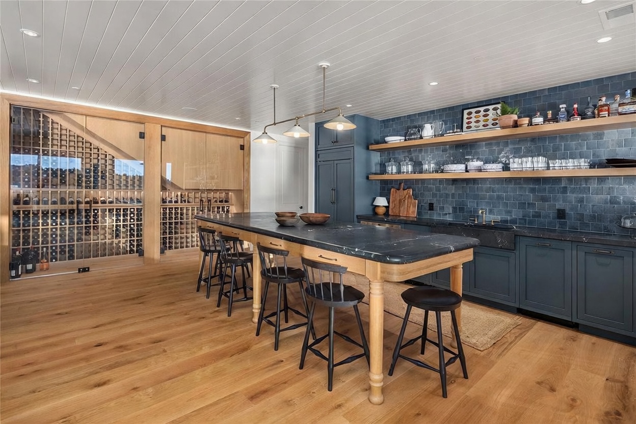 This sophisticated kitchen and bar area features a large central island with a dark stone countertop and light wood base, surrounded by black spindle-back stools. To the left, a glass-enclosed wine storage room showcases an extensive collection, while the right side features dark blue cabinetry, open wood shelving, and a dark stone sink. The space is illuminated by three pendant lights hanging from a white planked ceiling, creating a warm and inviting atmosphere.