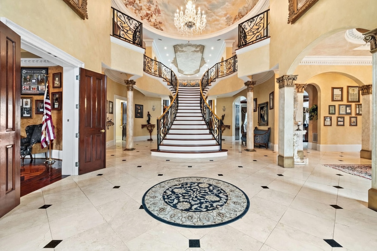 This grand foyer features a dramatic dual staircase with intricate wrought-iron railings that converge at a central landing. The space is defined by a high, vaulted ceiling with a painted mural and a large crystal chandelier, complemented by light-colored stone flooring with dark accent tiles. The perspective is centered, emphasizing the symmetrical architectural design and the opulent, formal aesthetic of the entryway.