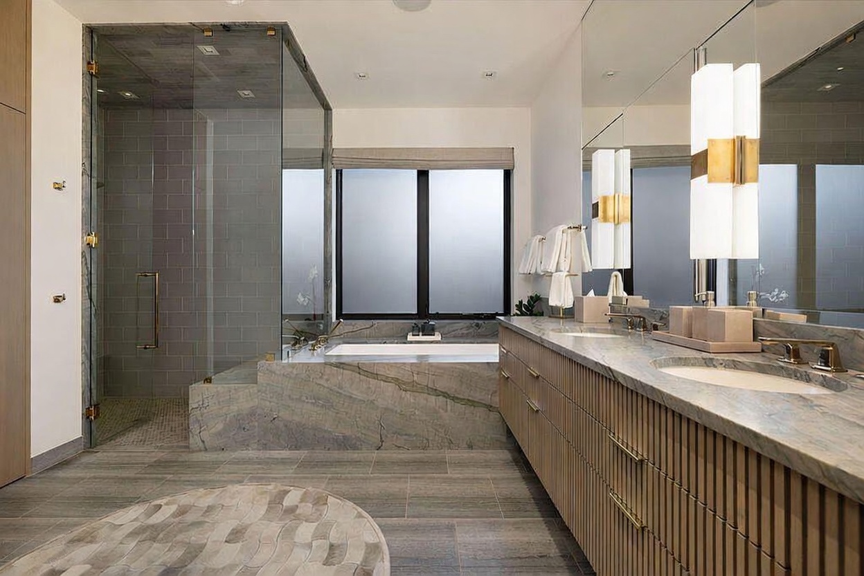 This luxurious primary bathroom features a large soaking tub encased in stone, positioned beneath a window with frosted glass. To the right, a long vanity with dual sinks and light wood cabinetry is topped with stone counters and illuminated by modern vertical light fixtures. A glass-enclosed shower with stone tile walls sits to the left, creating a clean, spa-like atmosphere with a sophisticated, contemporary aesthetic.