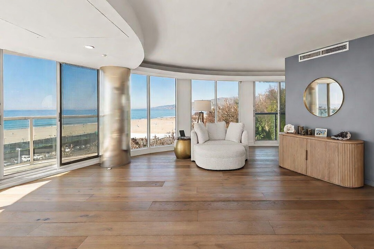 This bright and airy living space features expansive floor-to-ceiling windows that offer panoramic views of the beach and ocean. The room is anchored by a plush, curved white armchair and a light wood credenza, set against a soft gray accent wall with a circular mirror. The warm-toned wood flooring and minimalist decor create a serene, sophisticated atmosphere perfect for coastal living.