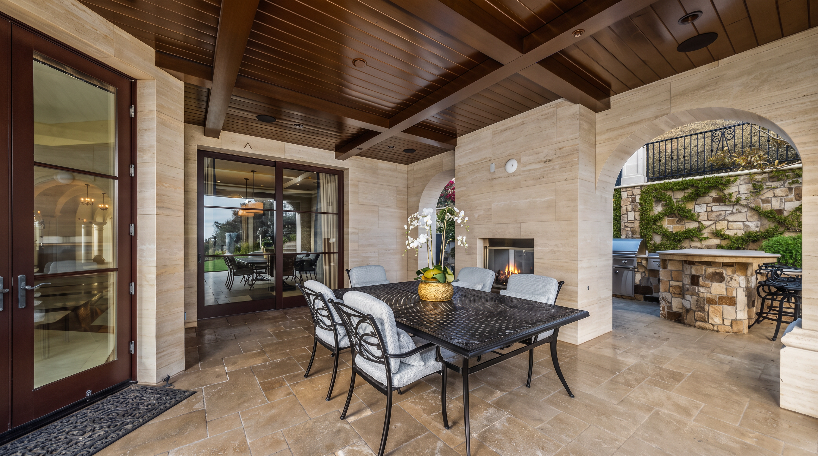 This covered outdoor patio features a dining set with a metal table and cushioned chairs, positioned in front of a stone-clad fireplace. The space is defined by a dark wood-paneled ceiling and light-toned stone walls, with a built-in outdoor kitchen area visible to the right. Large glass sliding doors provide a seamless transition to the interior, while the stone-paved floor and arched architectural details create an elegant, Mediterranean-inspired atmosphere.