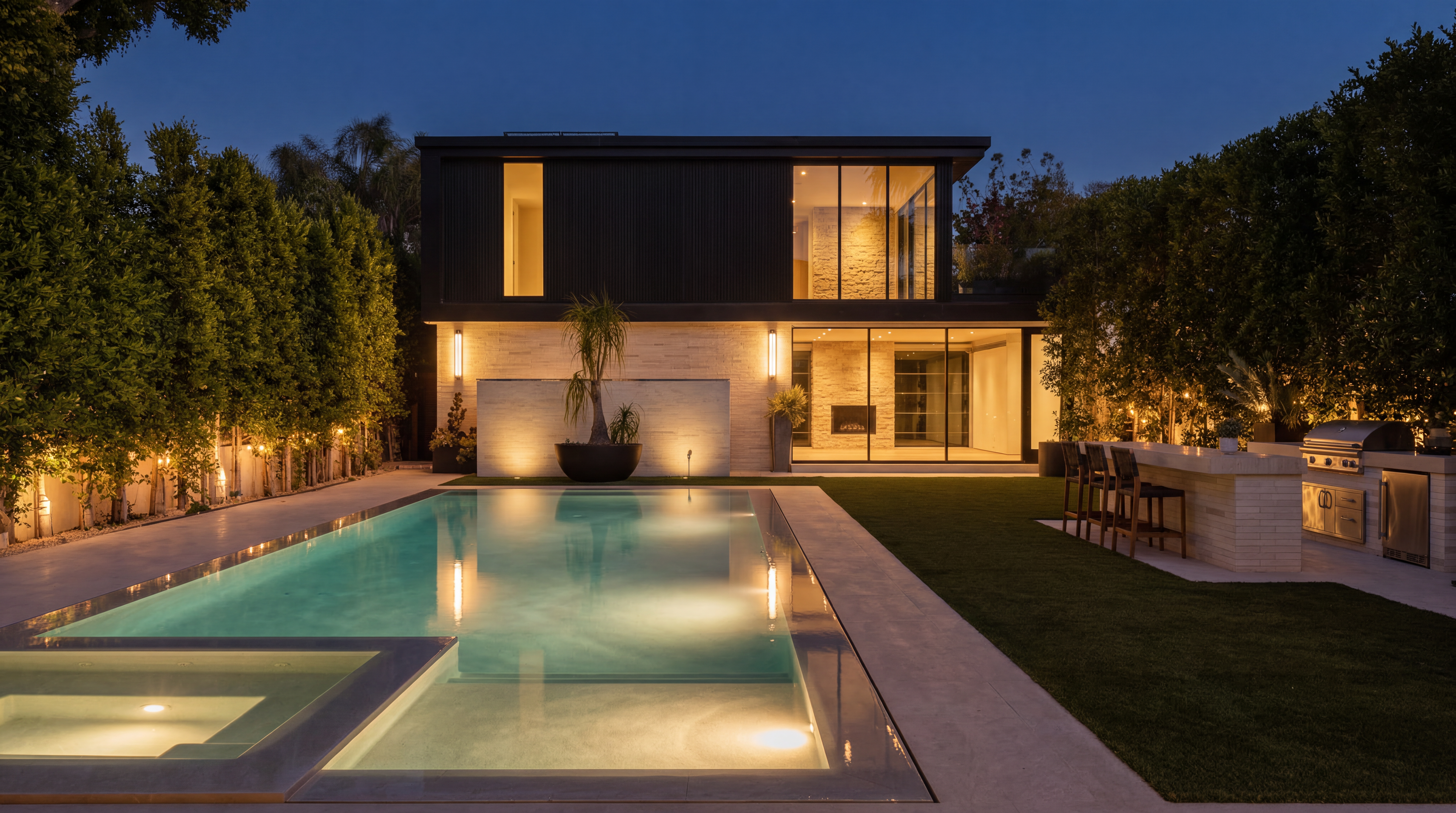 This modern, luxury residence showcases a stunning rear exterior design featuring a sleek dark-clad upper level contrasted by a textured light stone ground floor. The expansive backyard includes a lit swimming pool and spa adjacent to a well-manicured lawn and a sophisticated outdoor kitchen with bar seating. The atmospheric evening lighting highlights the contemporary architecture and creates an inviting, high-end outdoor oasis perfect for entertaining.
